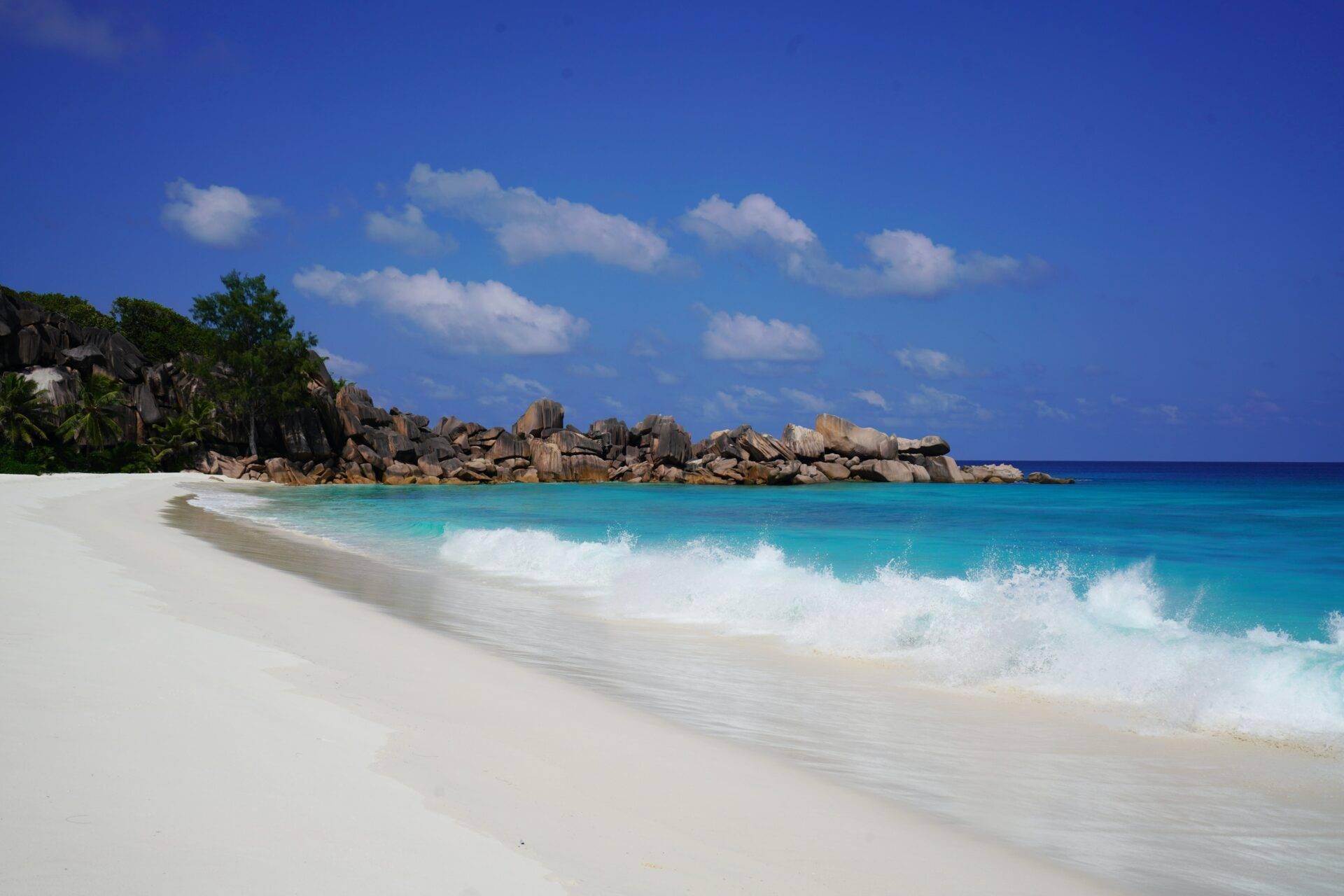 Plage de sable blanc avec vagues turquoise et formations rocheuses granitiques sous un ciel bleu dégagé