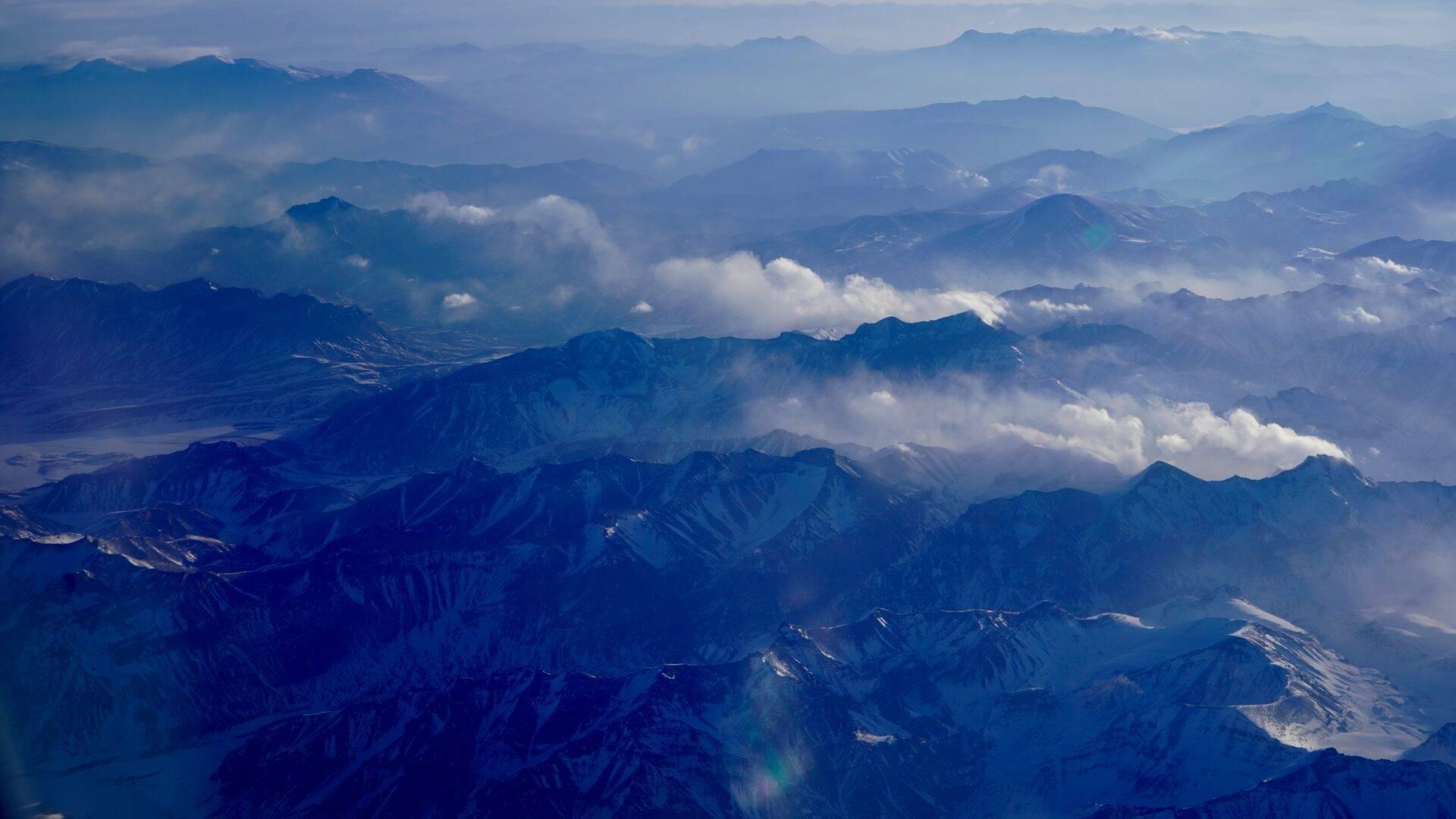 Aerial view of a major mountain range with snow-capped peaks, deep valleys and scattered white clouds