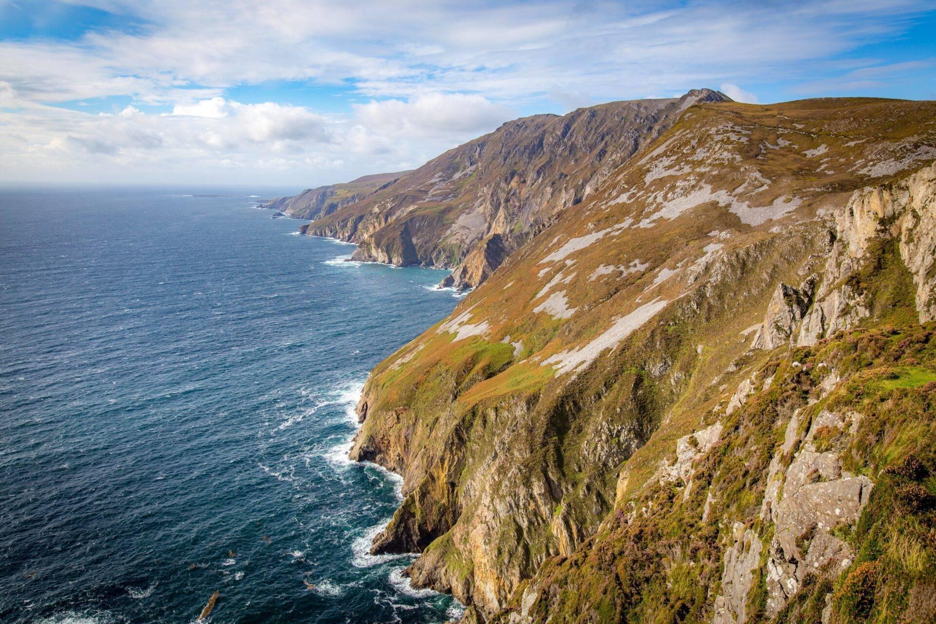 Steep green coastal cliffs rising above the Atlantic Ocean under a cloudy sky
