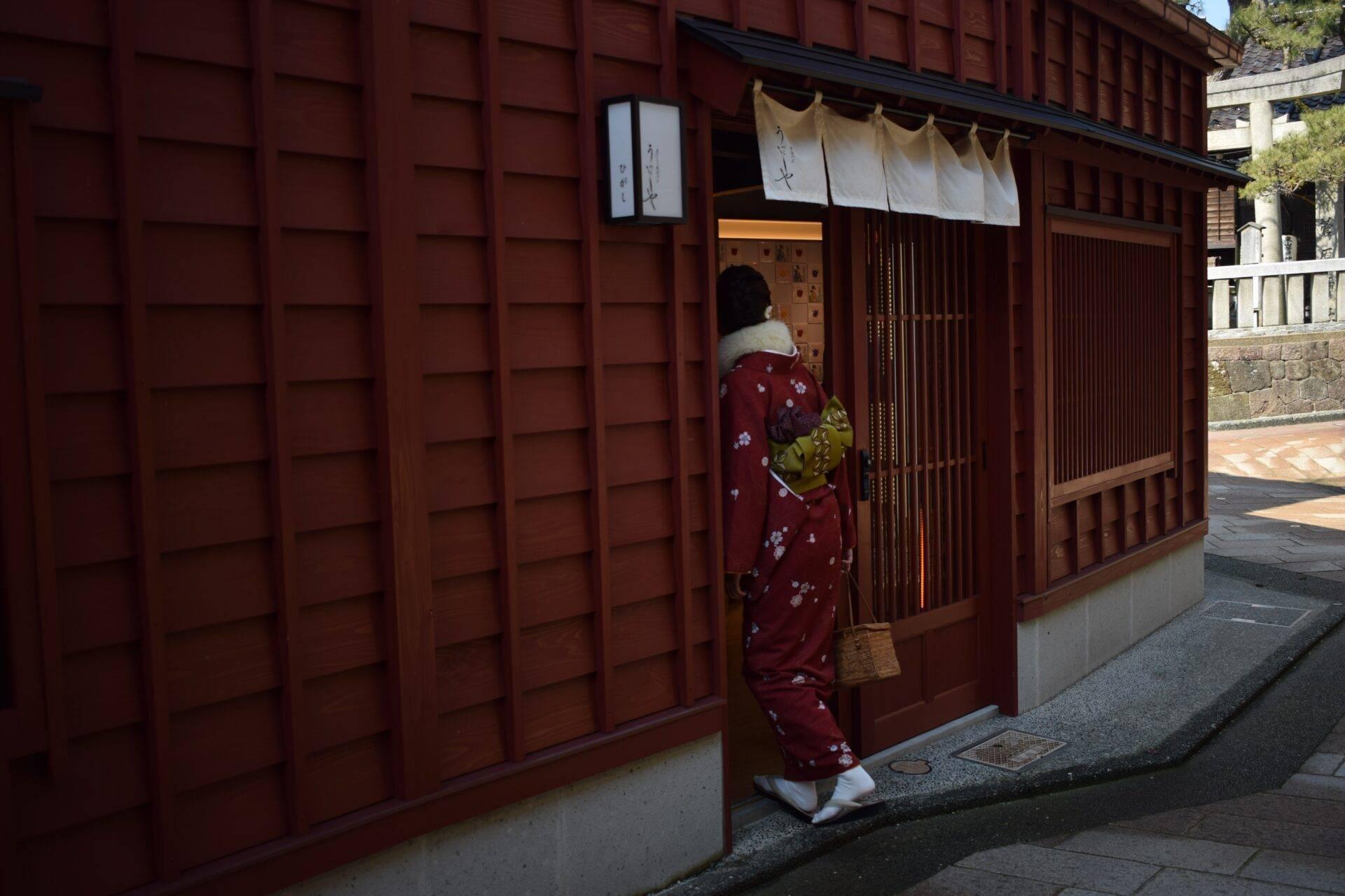 A woman in a red kimono stepping into a traditional Japanese establishment with dark wood walls and doors