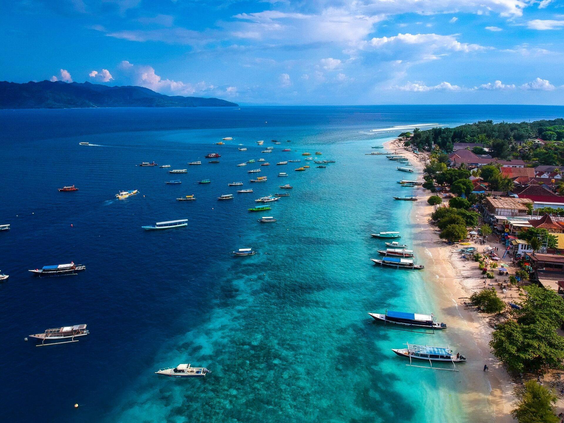 Aerial view of a tropical bay with colourful sailing boats anchored near a white sand beach bordered by houses
