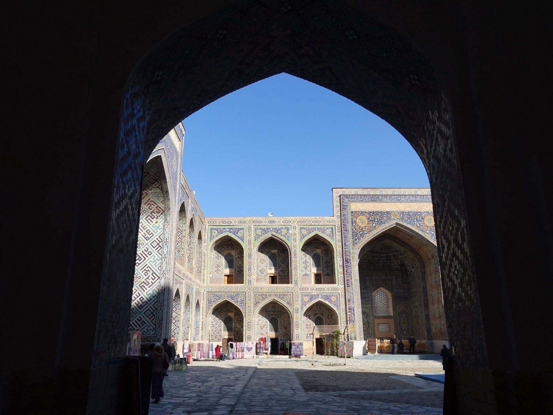 View through an entrance arch into a courtyard adorned with blue mosaic tilework and geometric Islamic patterns