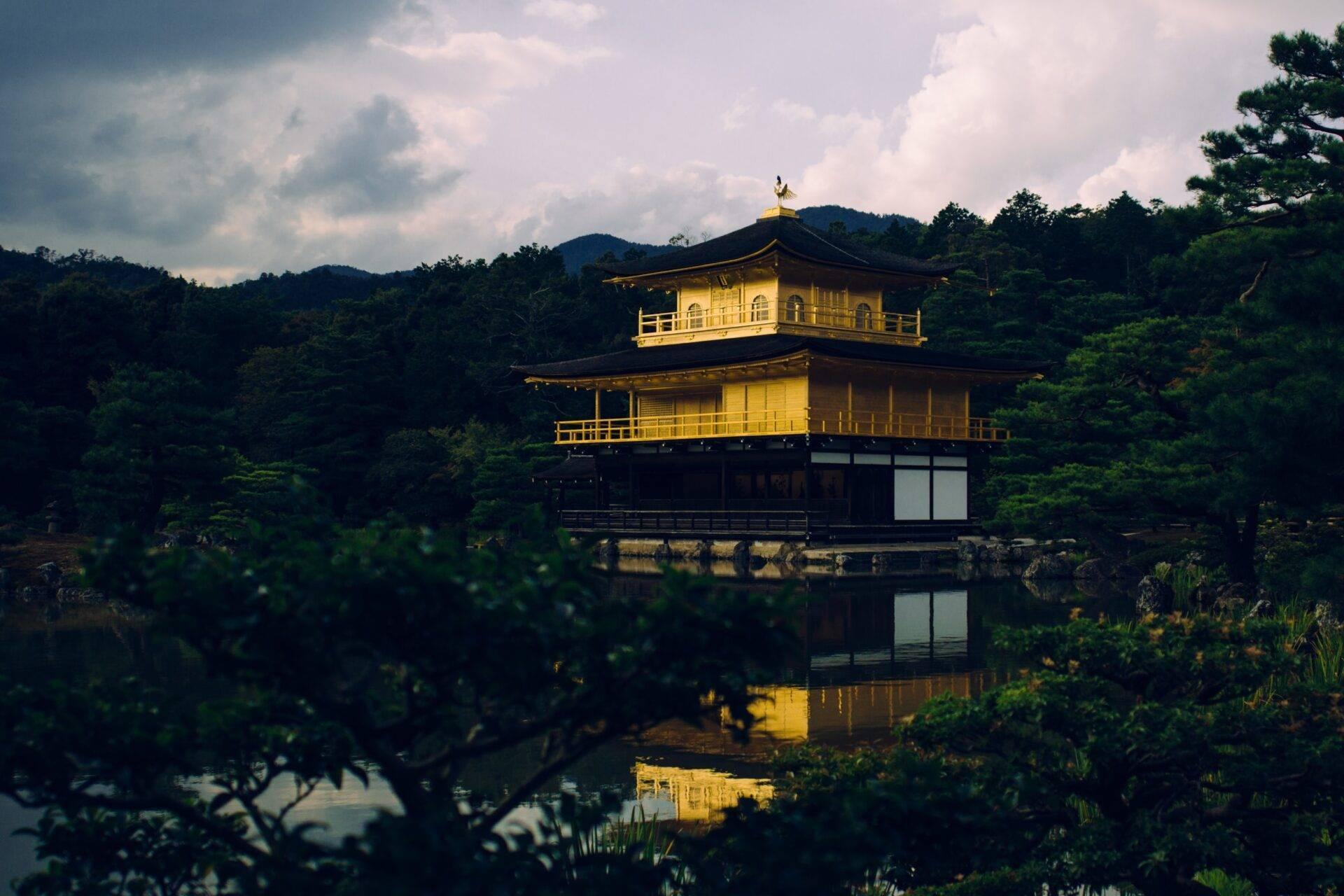 A two-storey golden pavilion reflected in a calm pond, surrounded by greenery and wooded hills