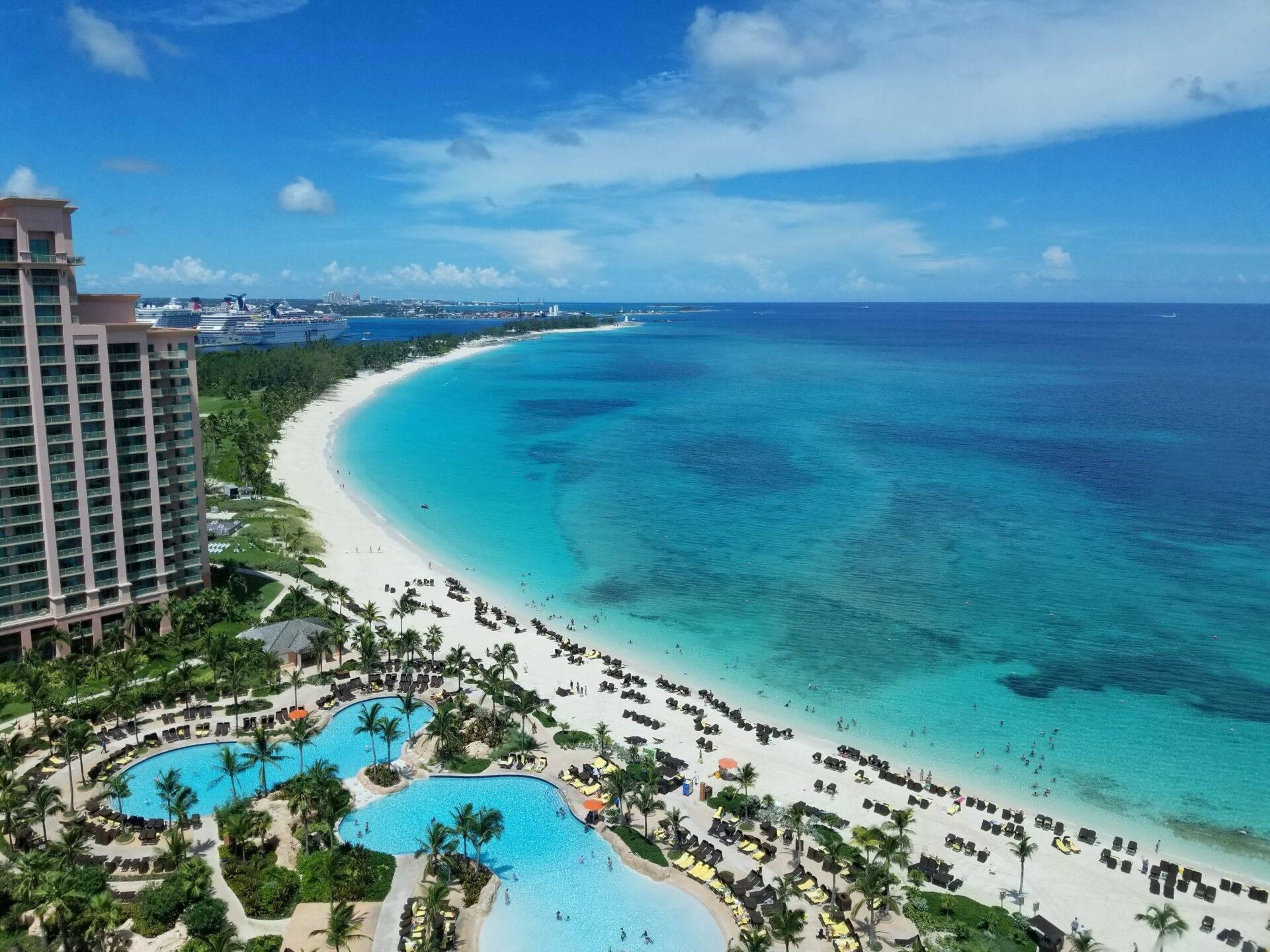 Vue aérienne d’une station balnéaire avec plage de sable blanc, piscines turquoise et océan bleu tropical parsemé de navires de croisière
