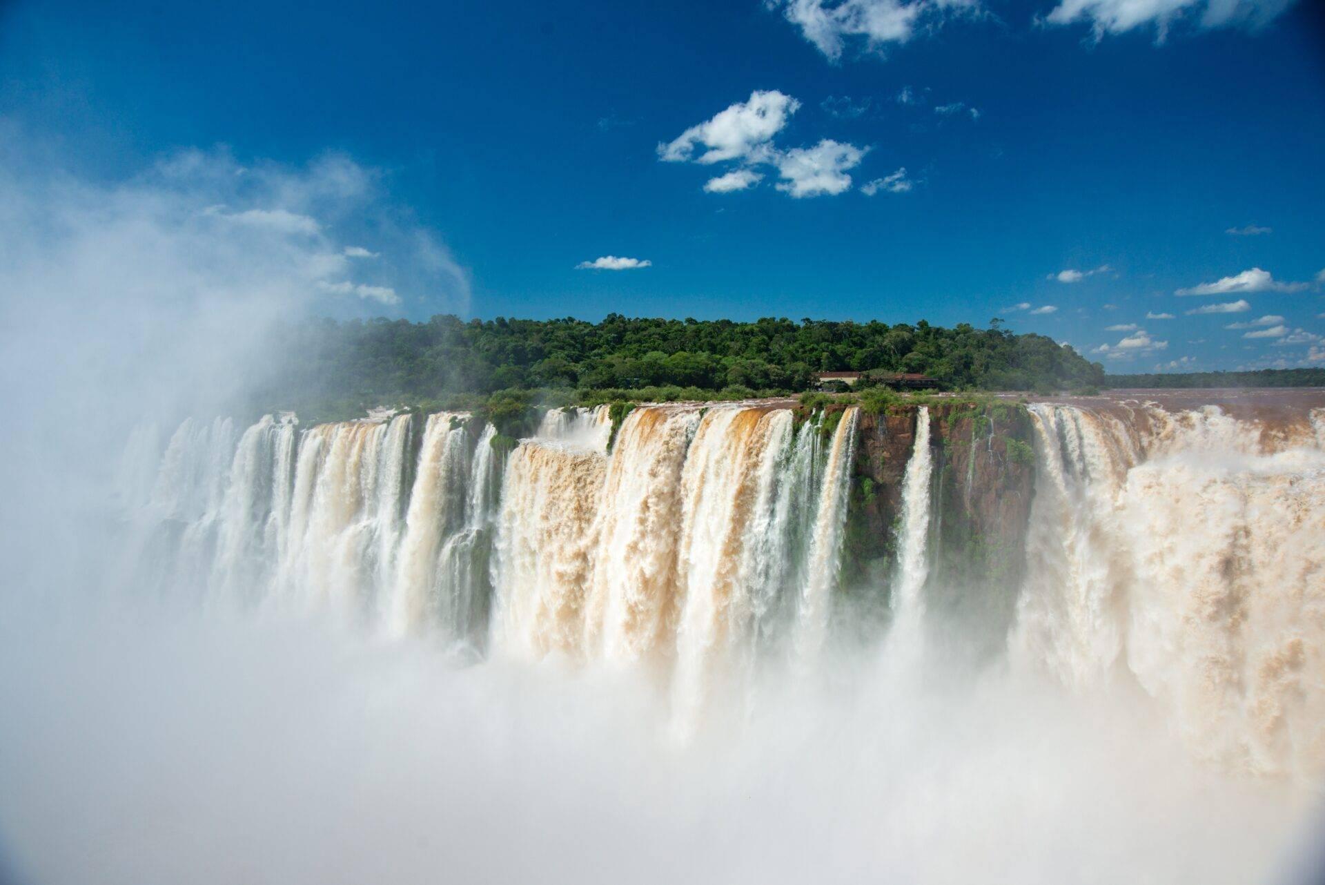 Powerful waterfalls cascading in thick white mist, framed by lush tropical forest and blue sky
