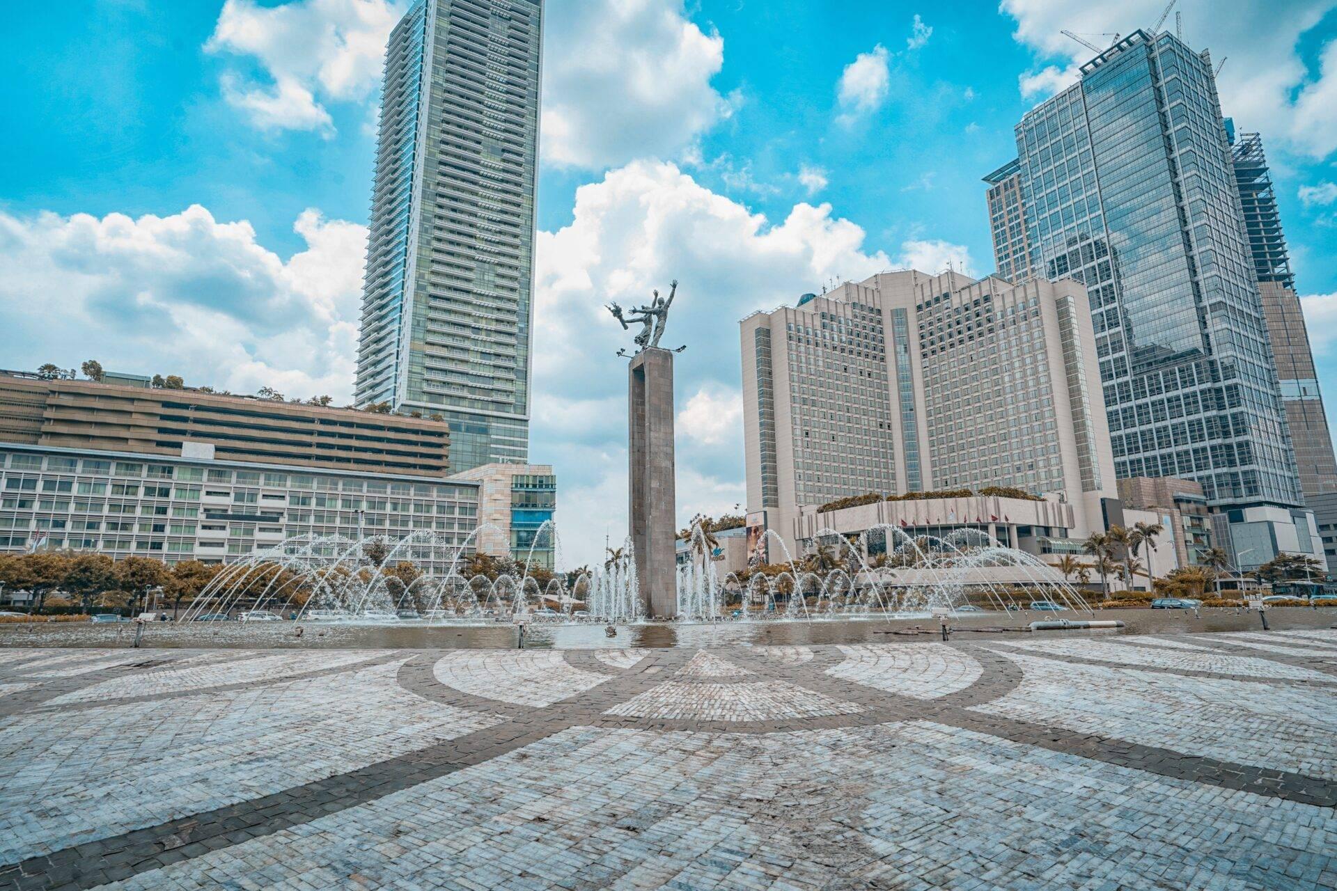 A paved urban square with a monumental statue, fountains and modern skyscrapers under a blue sky