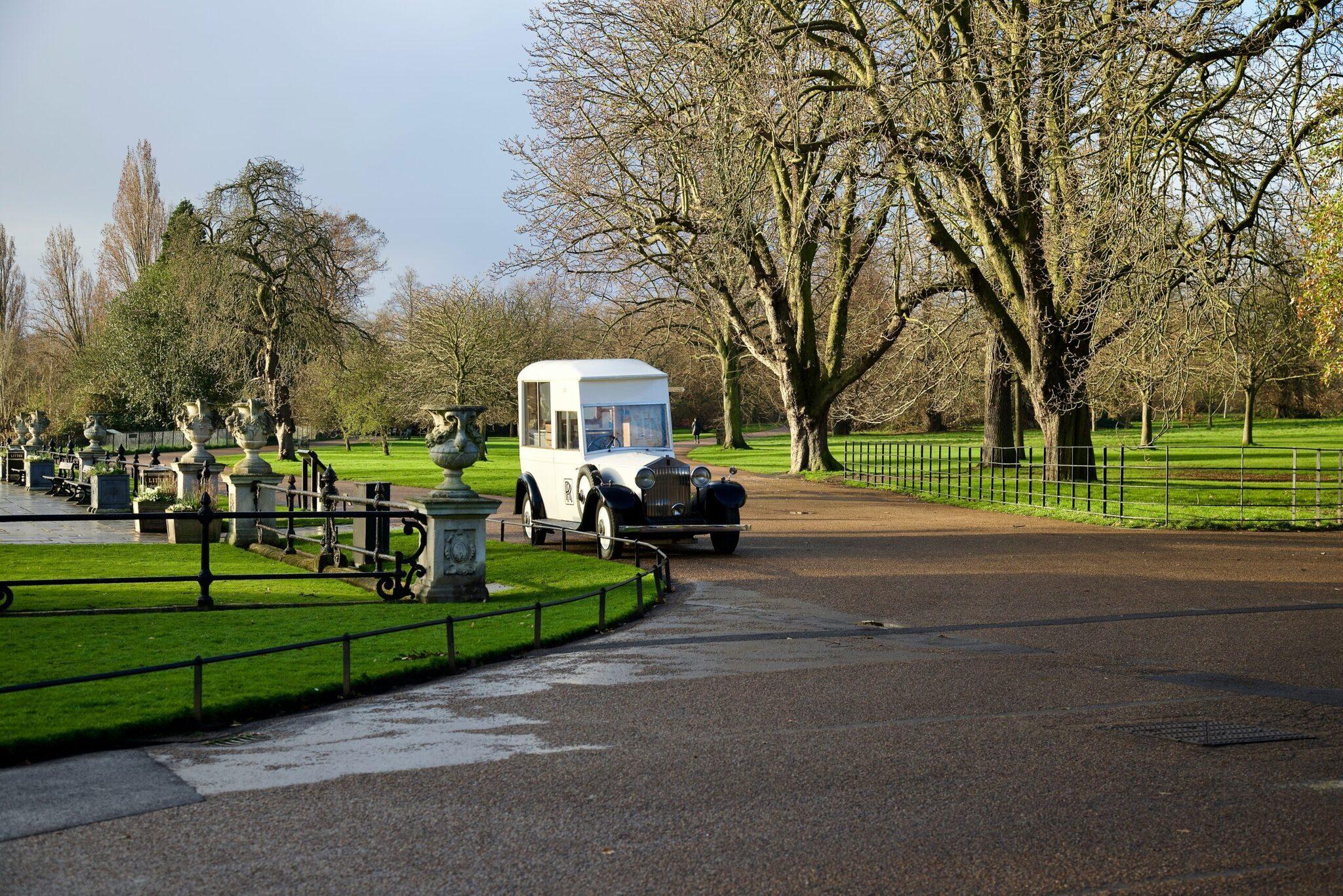 Une voiture vintage de mariage blanche et noire circule dans un parc urbain verdoyant bordé d’arbres centenaires