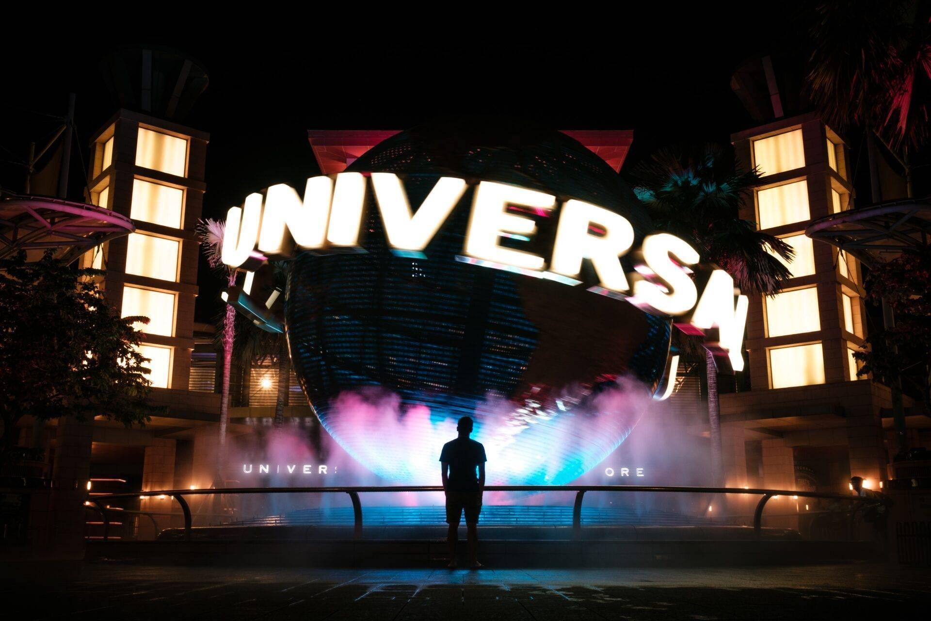 Un visiteur face à l’entrée illuminée d’un parc d’attractions Universal, de nuit, avec globe géant et effets lumineux colorés