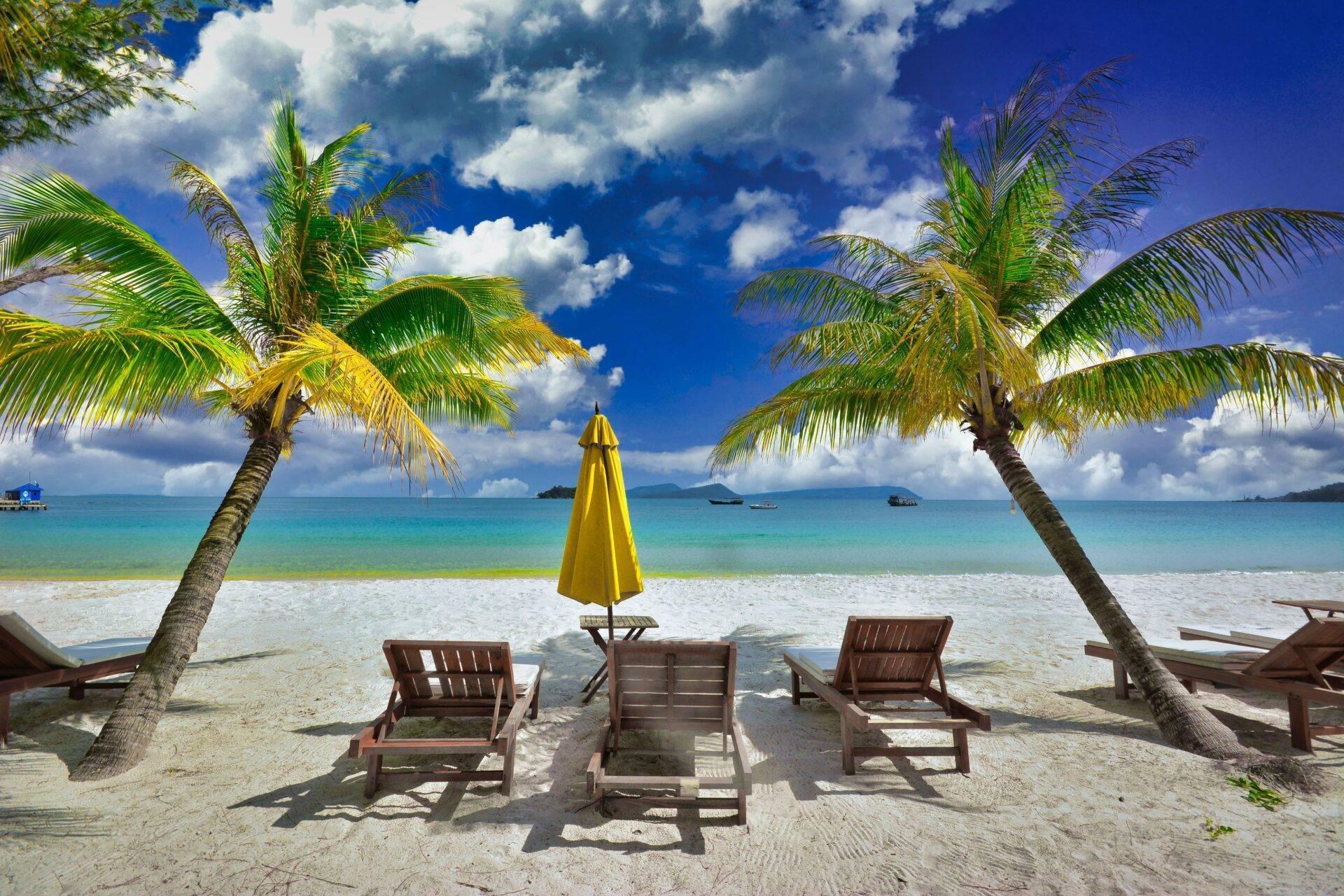 Plage tropicale avec transats en bois, parasol jaune et palmiers encadrant une mer turquoise sous ciel bleu nuageux