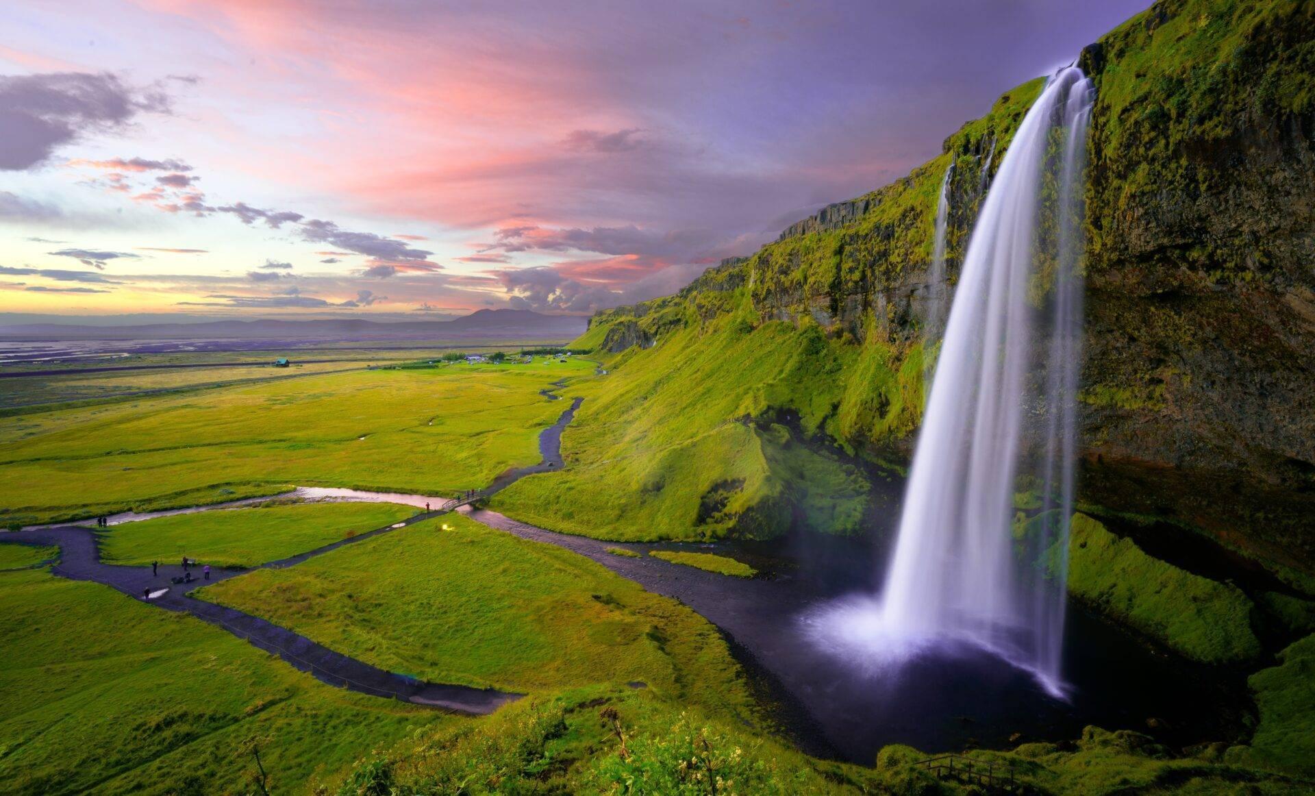 A spectacular waterfall cascades over a green clifftop overlooking a river valley at sunset