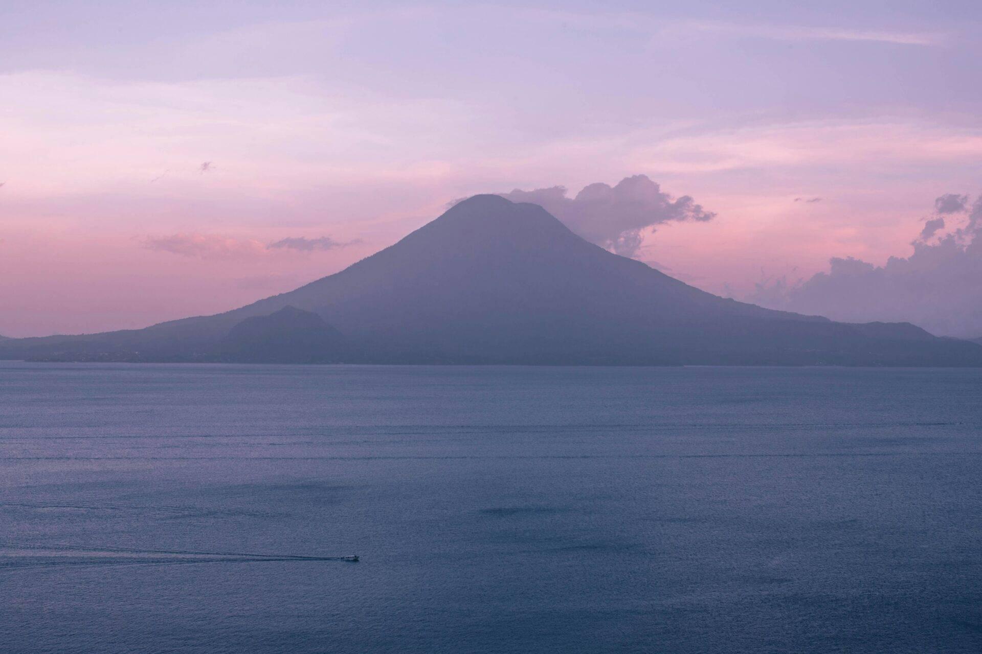 Volcan aux pentes régulières dominant un lac calme au coucher du soleil dans une atmosphère rose et violette