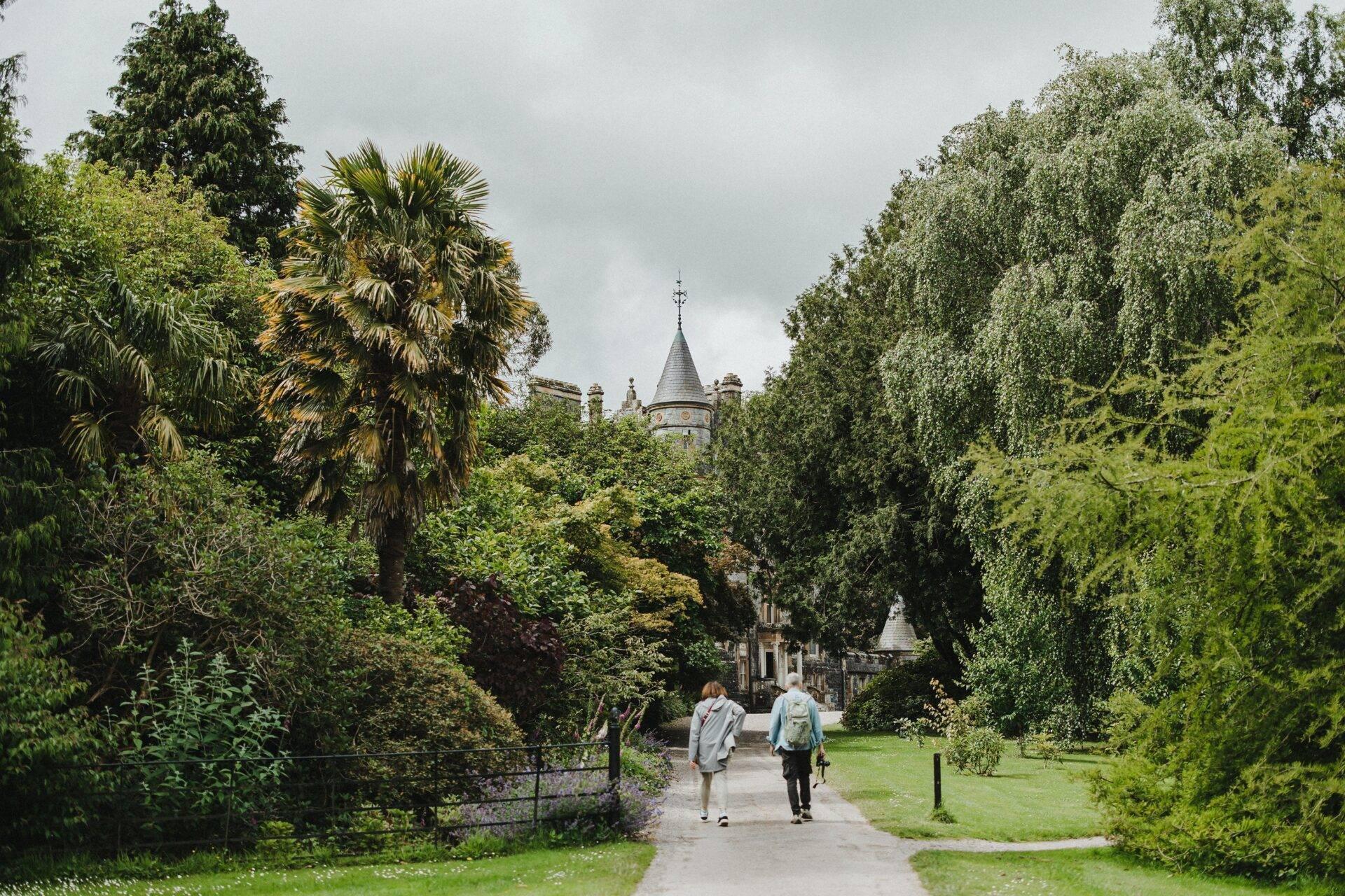 Two hikers walking along a shaded path towards a castle with a pointed tower surrounded by greenery