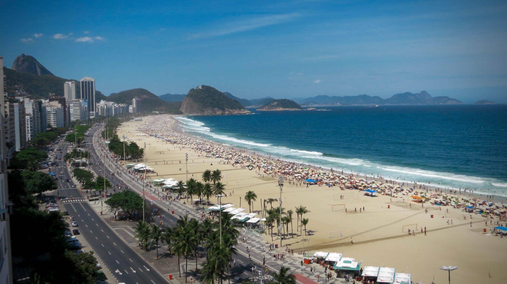 Aerial view of a crowded urban beach with city buildings in the background, lush green mountains and blue ocean