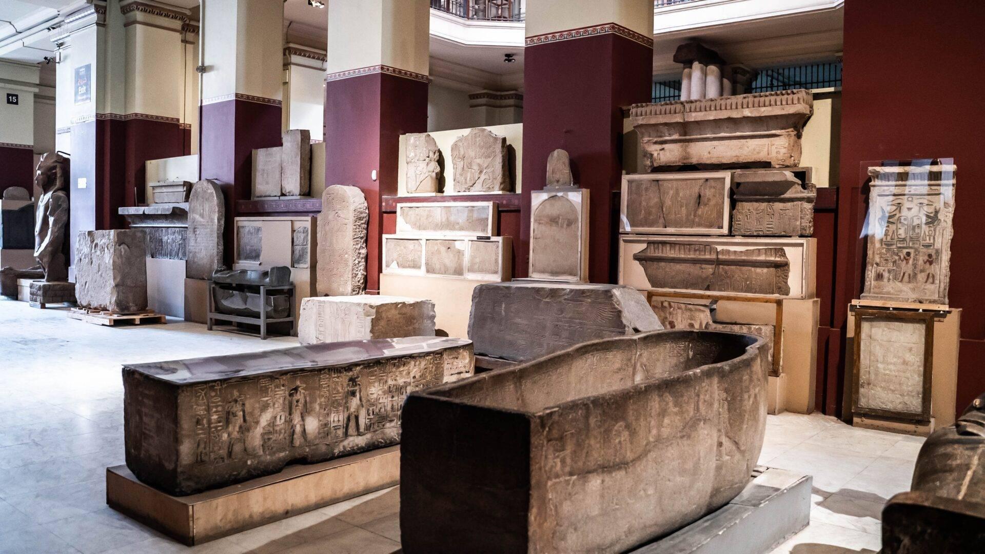 Ancient Egyptian sarcophagi and bas-reliefs displayed in a museum gallery with deep red walls