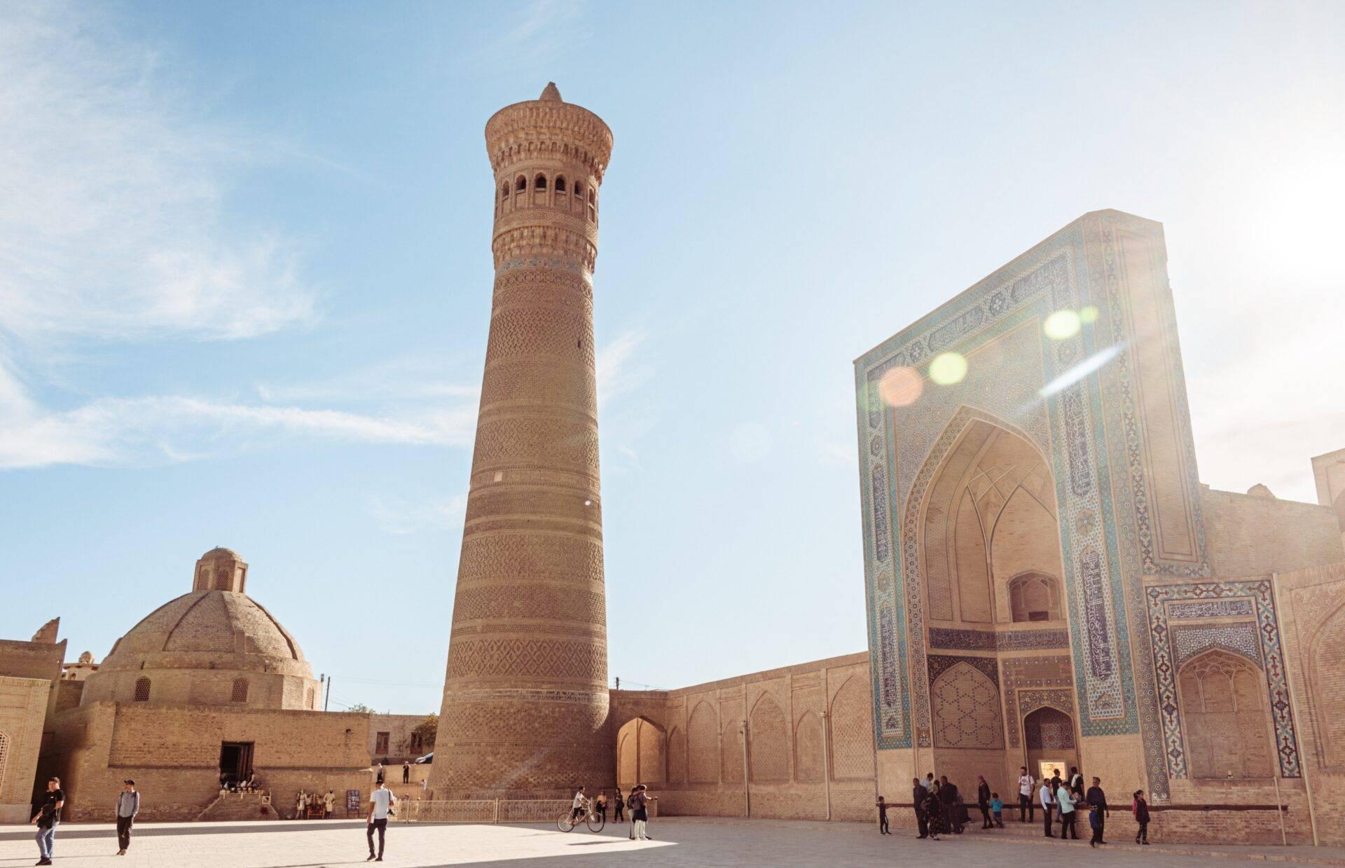 Historic madrasa courtyard with a brick minaret, monumental ornamental gateway and visitors under a clear sky