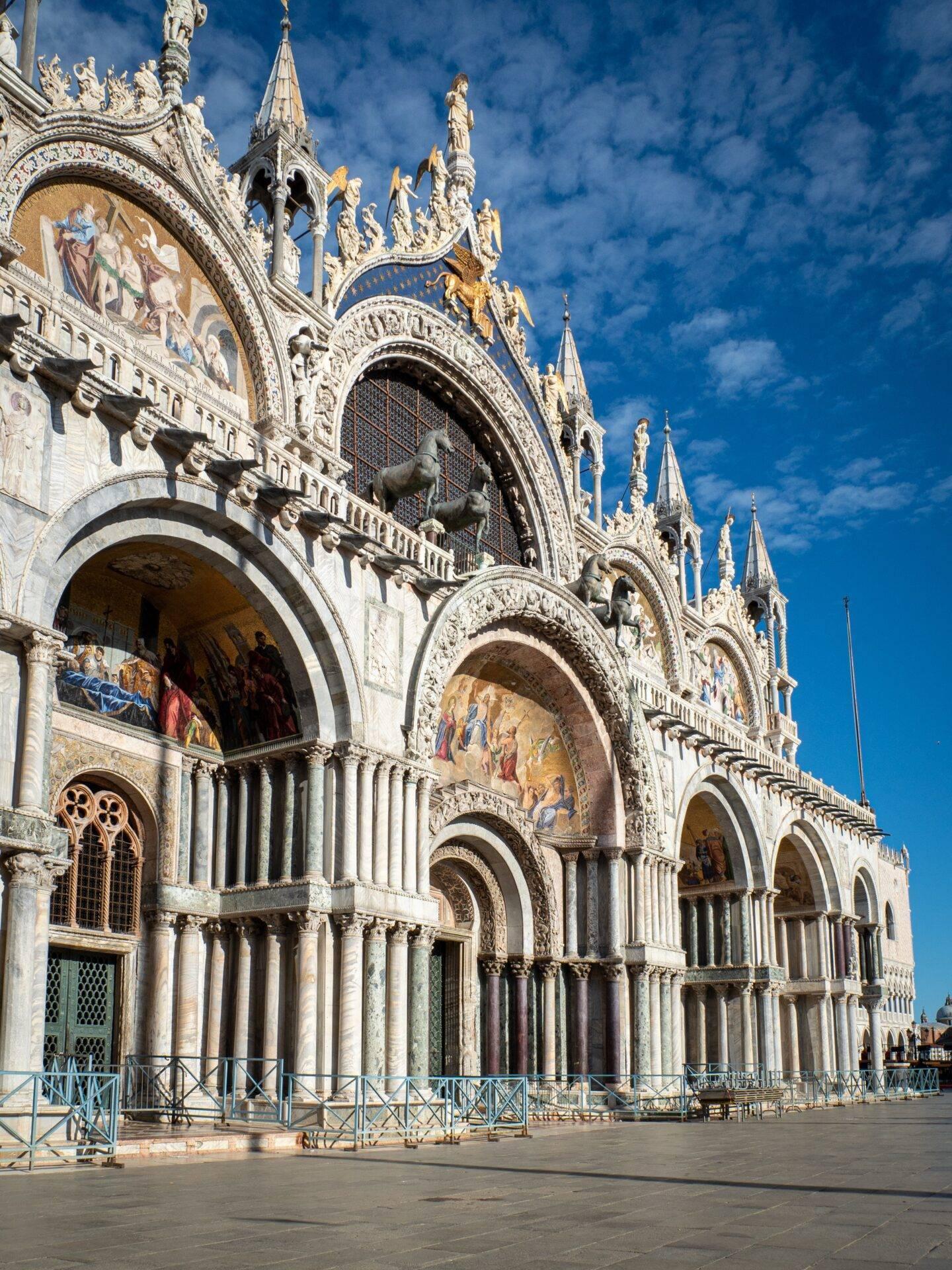 Ornate facade of a Venetian basilica with Gothic arcades, gilded sculptures and mosaics under a blue sky