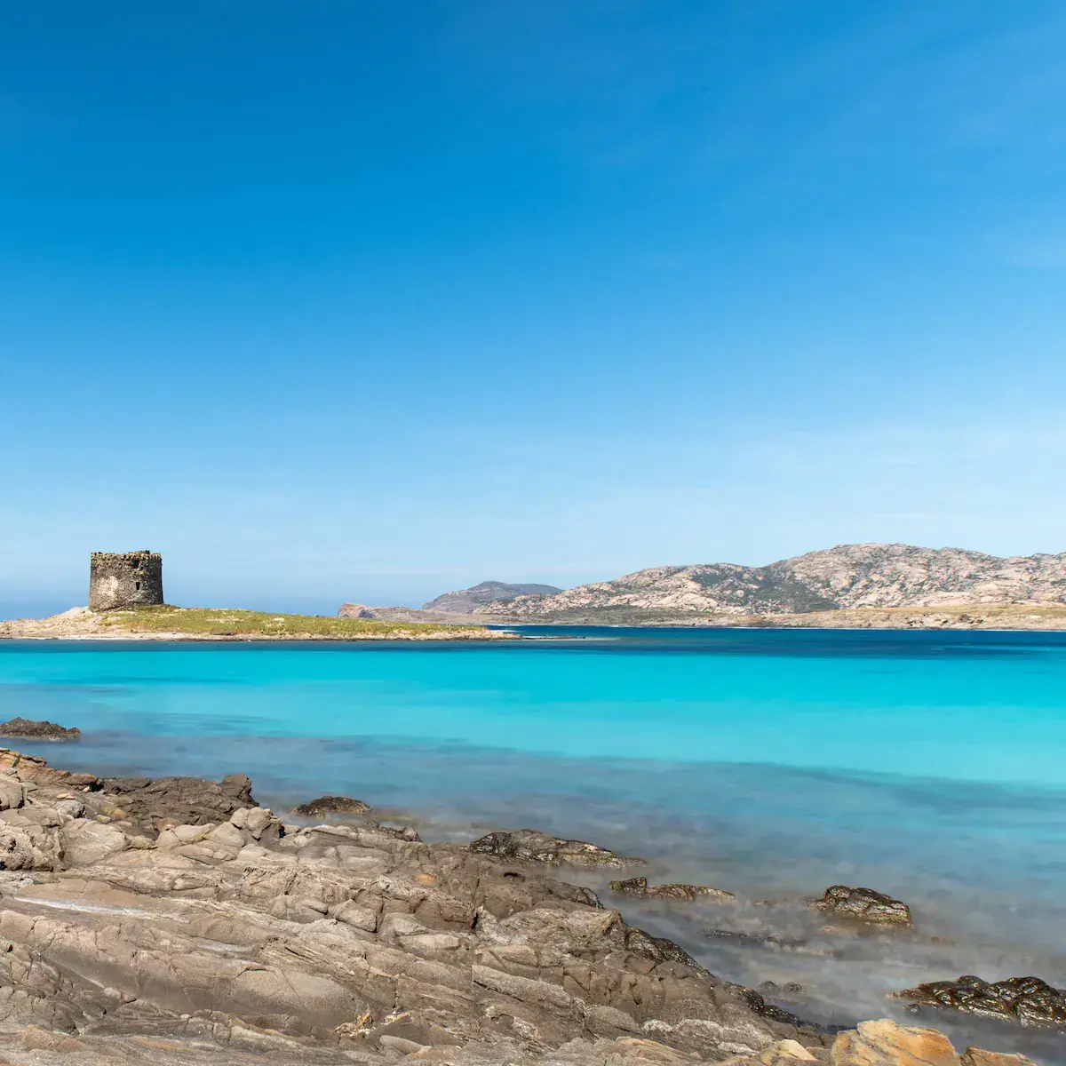 Une tour de pierre ancienne se dresse sur une côte rocheuse face à une mer turquoise et des collines arides