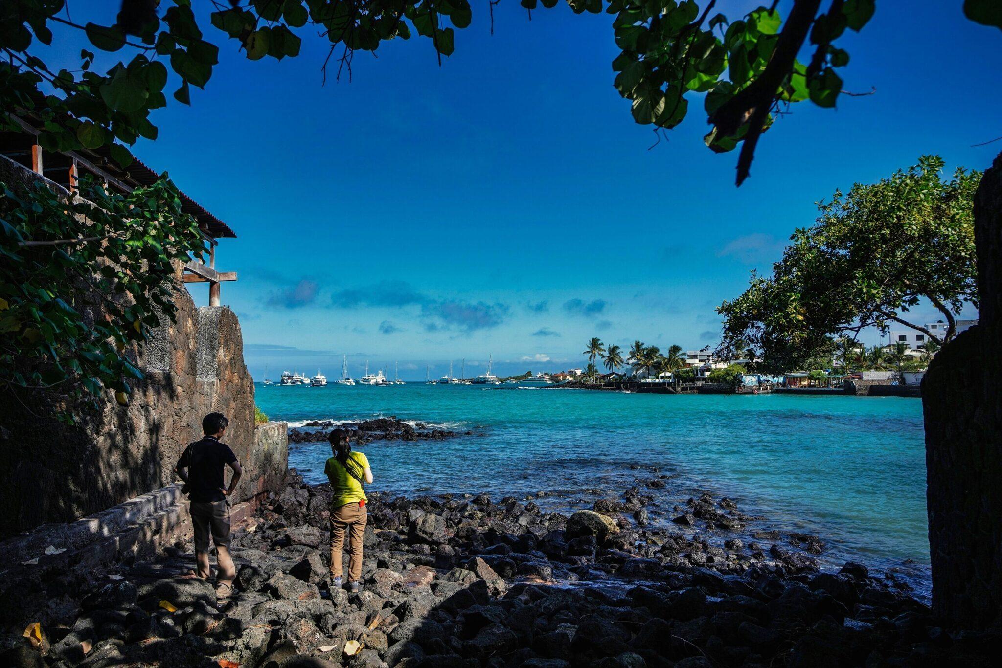 Deux randonneurs observent une baie tropicale turquoise avec voiliers depuis des rochers près d’un temple côtier