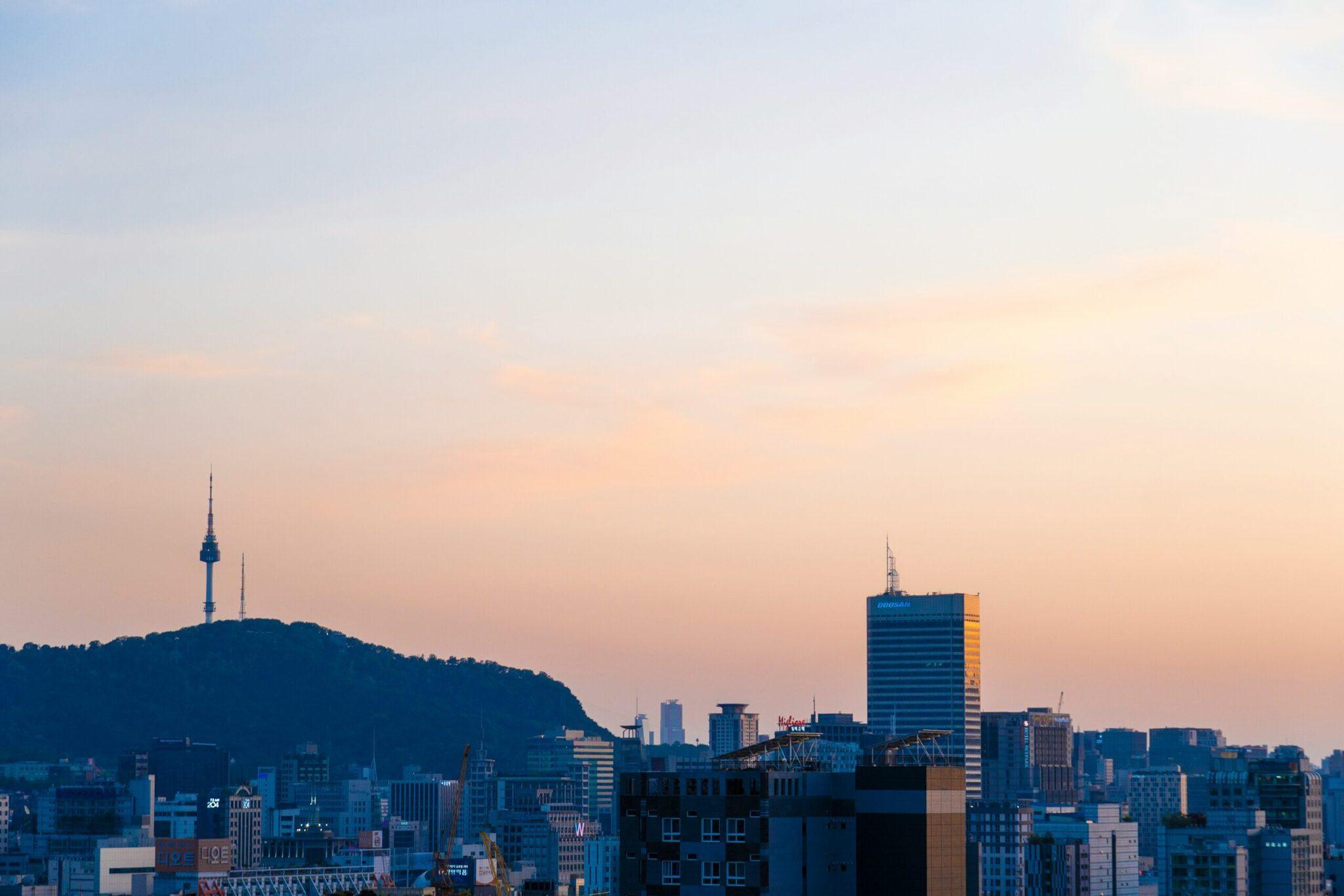 City skyline at sunset with a communications tower on a wooded hill and modern skyscrapers in the background