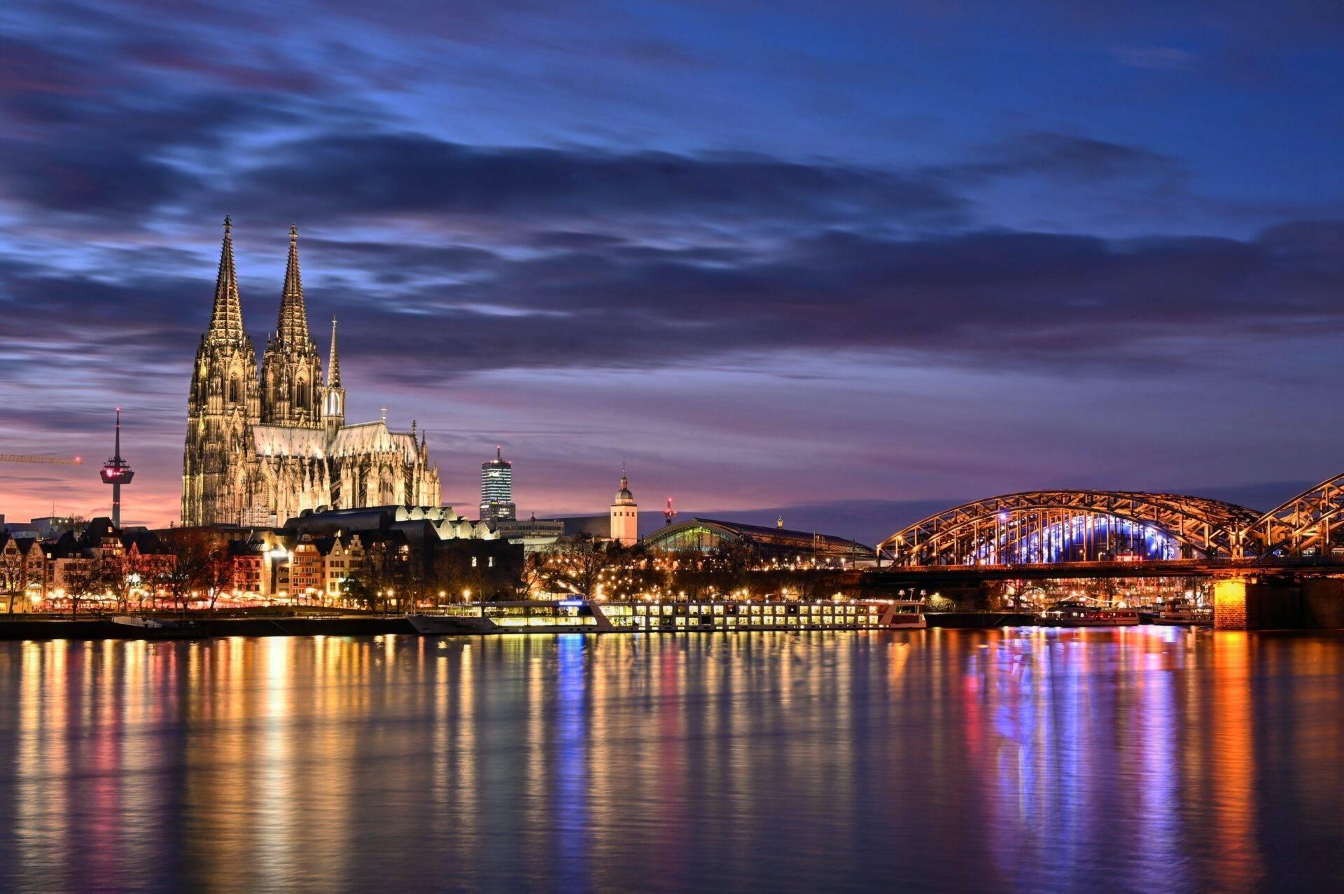 Gothic cathedral illuminated at dusk with a metal bridge and reflections shimmering in the river below