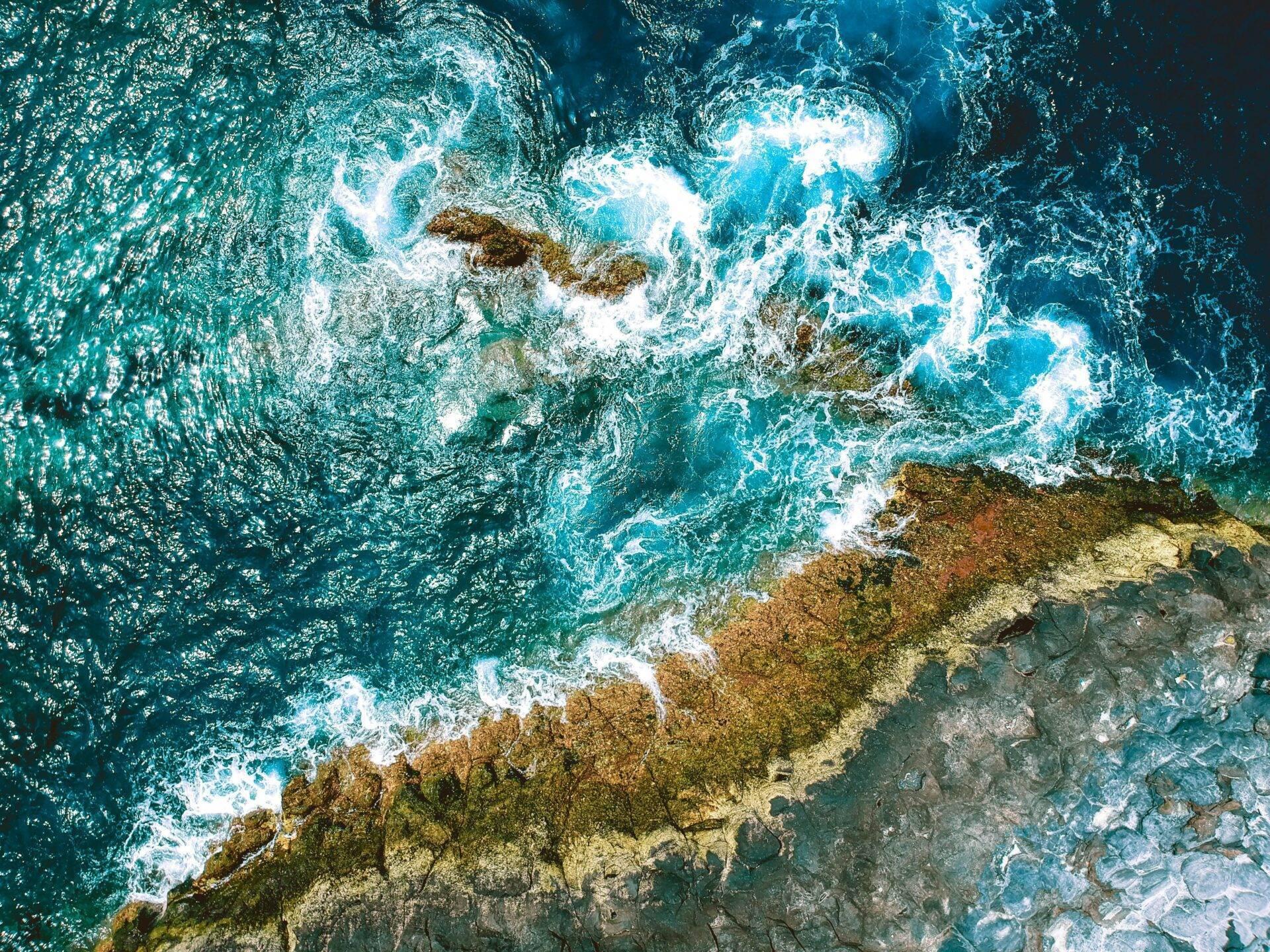 Vue aérienne de vagues turquoise déferlant contre une falaise rocheuse côtière aux teintes dorées et grises