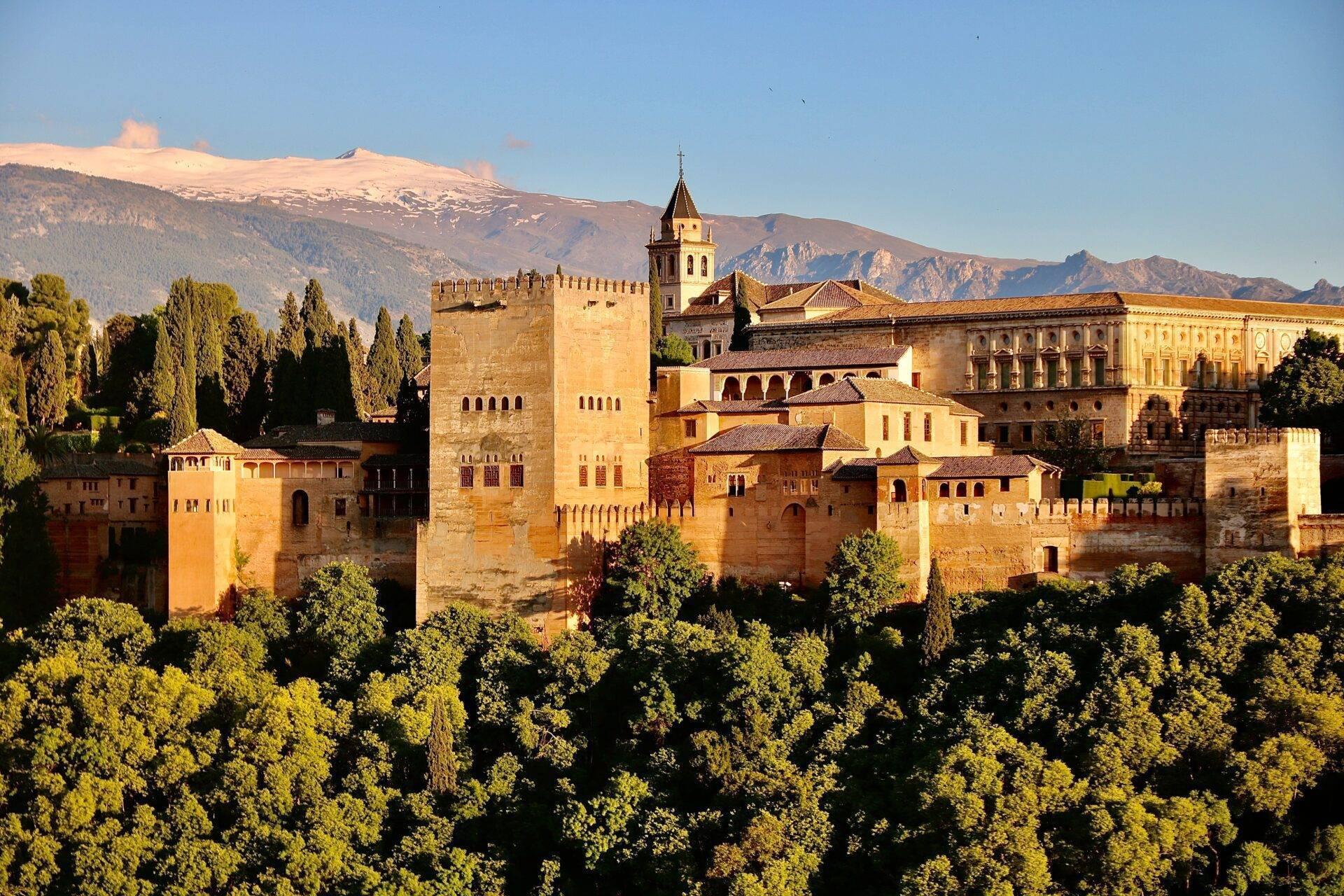 Forteresse médiévale dorée avec tours et murailles, entourée de végétation verdoyante et montagnes enneigées au loin