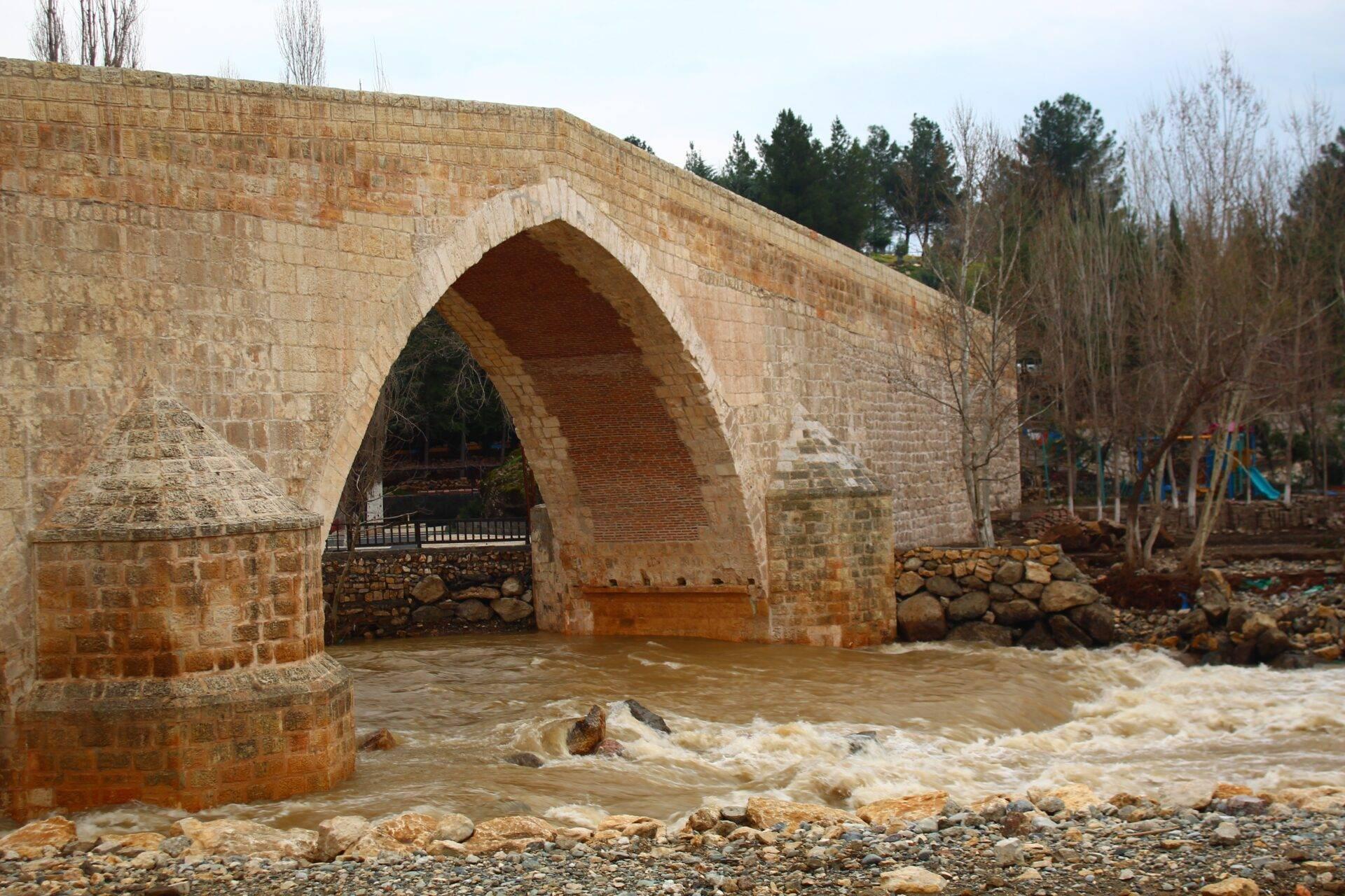 An ancient stone bridge with a wide arch spanning a river in flood with murky, fast-flowing water