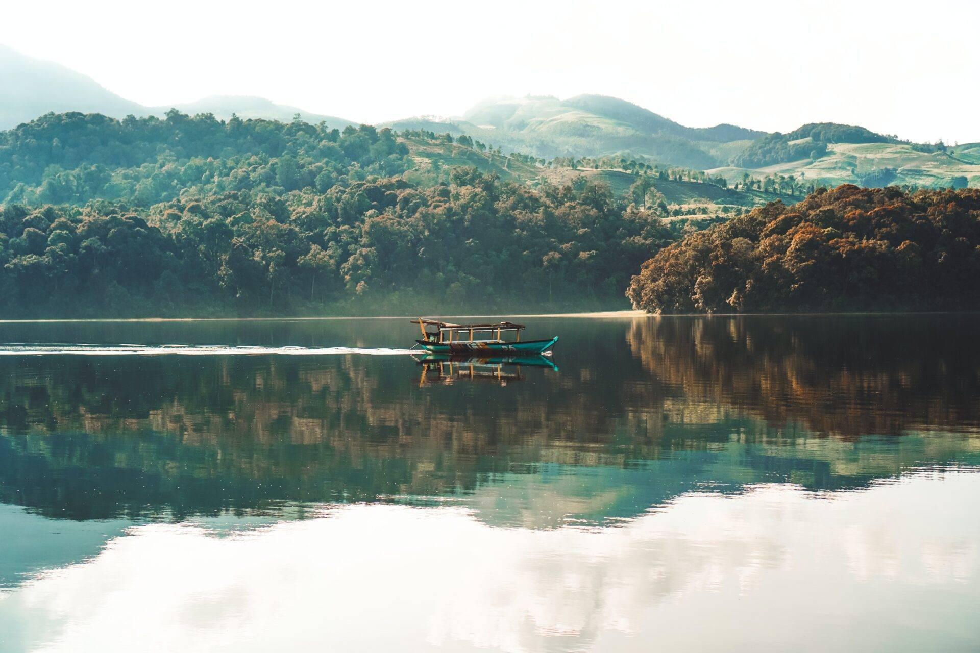 A traditional blue fishing boat on a mirror-calm lake surrounded by forested mountains and lush jungle