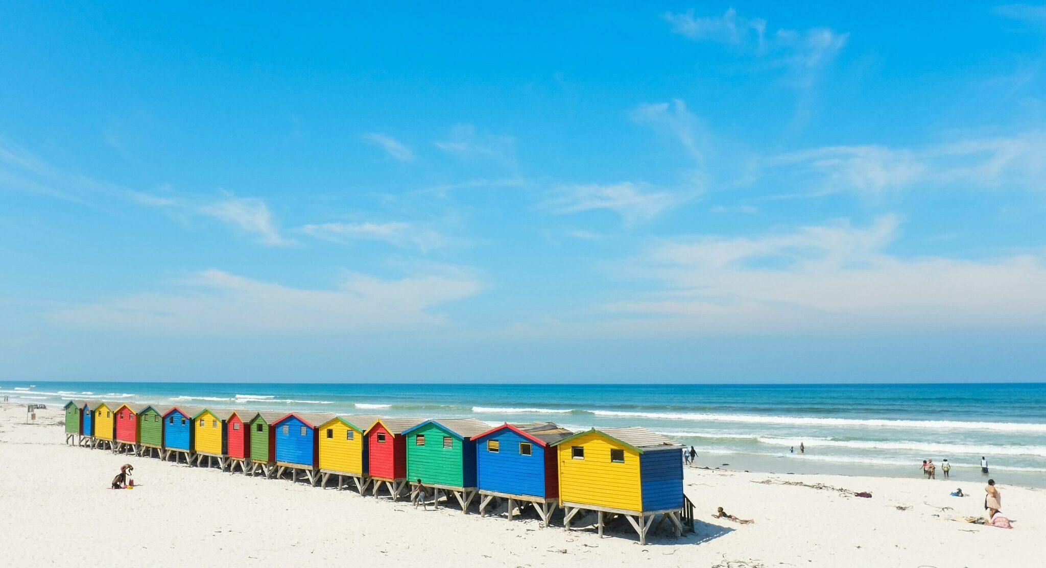 A row of colourful beach huts lined up on white sand facing the turquoise ocean