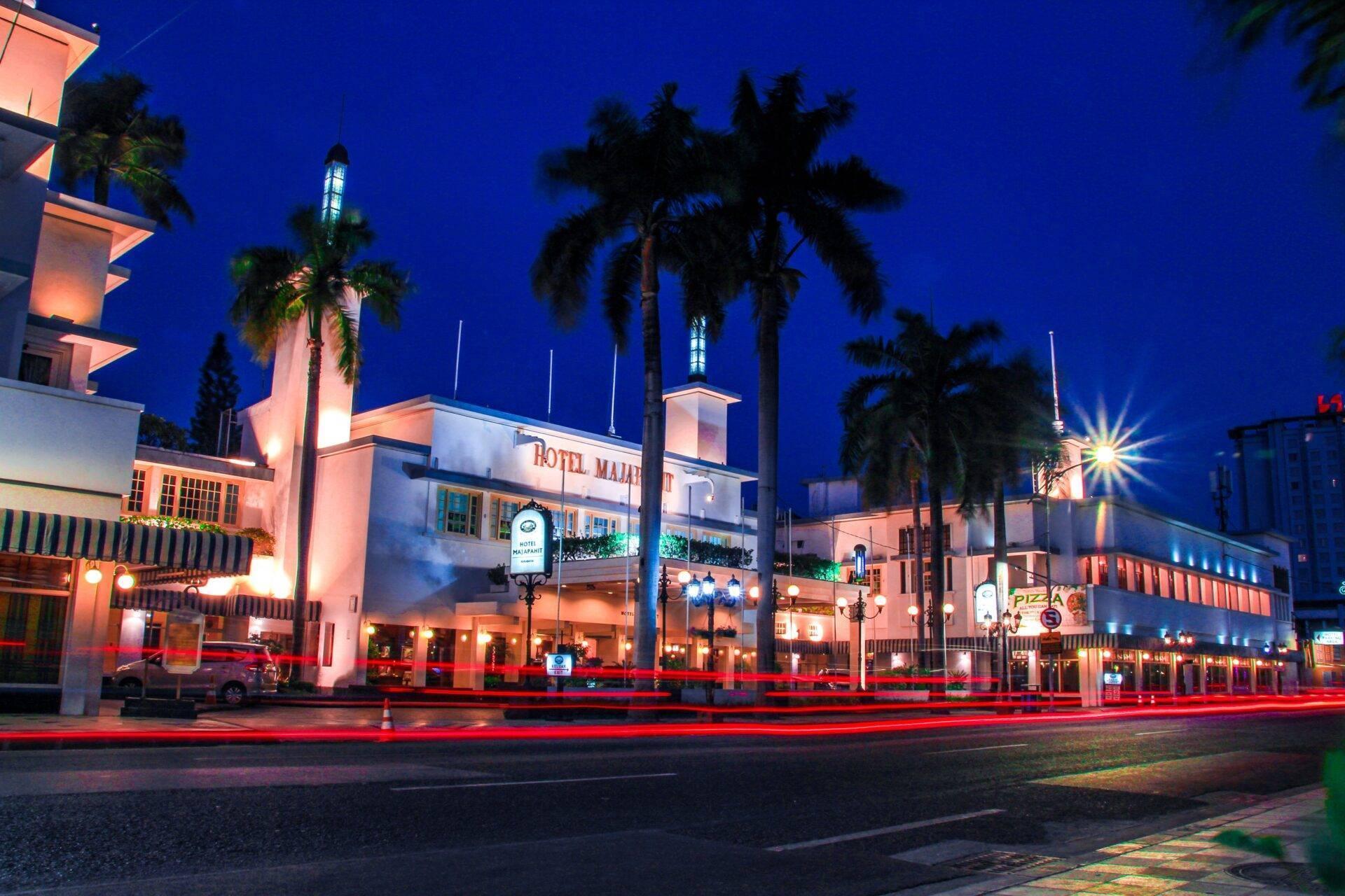 The illuminated facade of a colonial hotel at night with palm trees, Art Deco architecture and red light trails from passing traffic