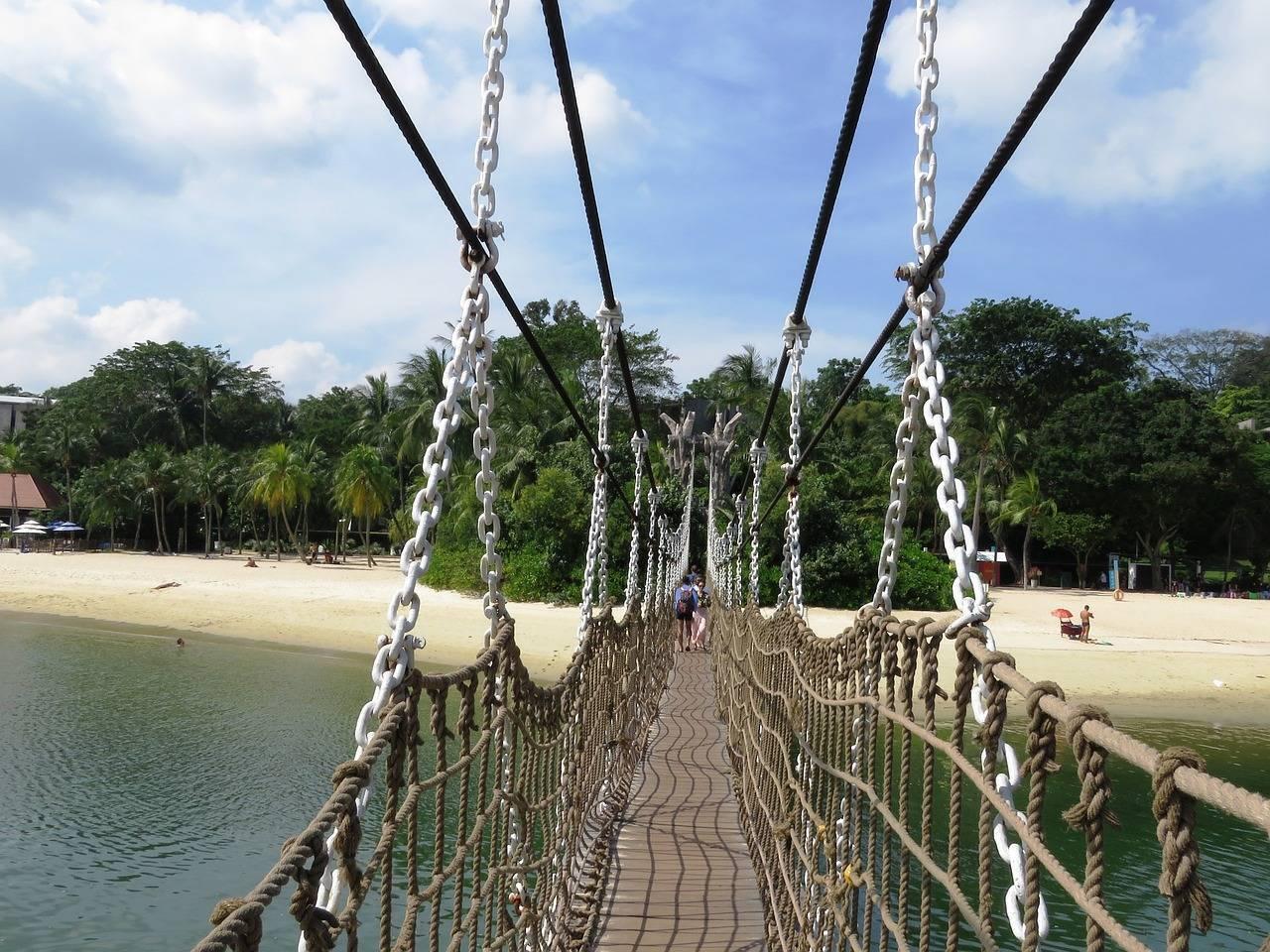 Un pont suspendu en cordes et chaînes métalliques surplombant une plage tropicale avec végétation luxuriante