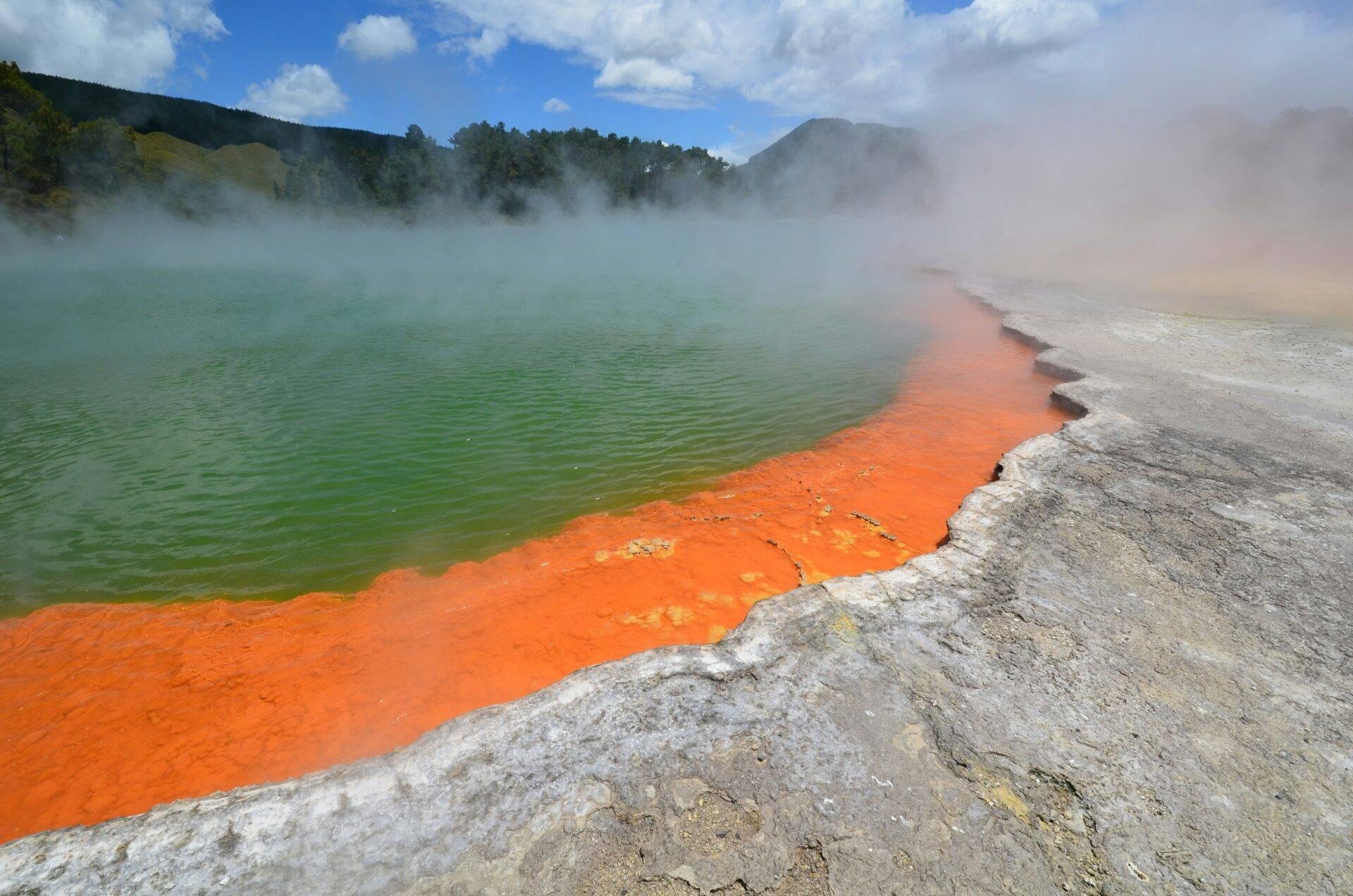 Un bassin géothermique aux eaux colorées vert émeraude et orange vif, entouré de vapeur et de formations minérales blanches