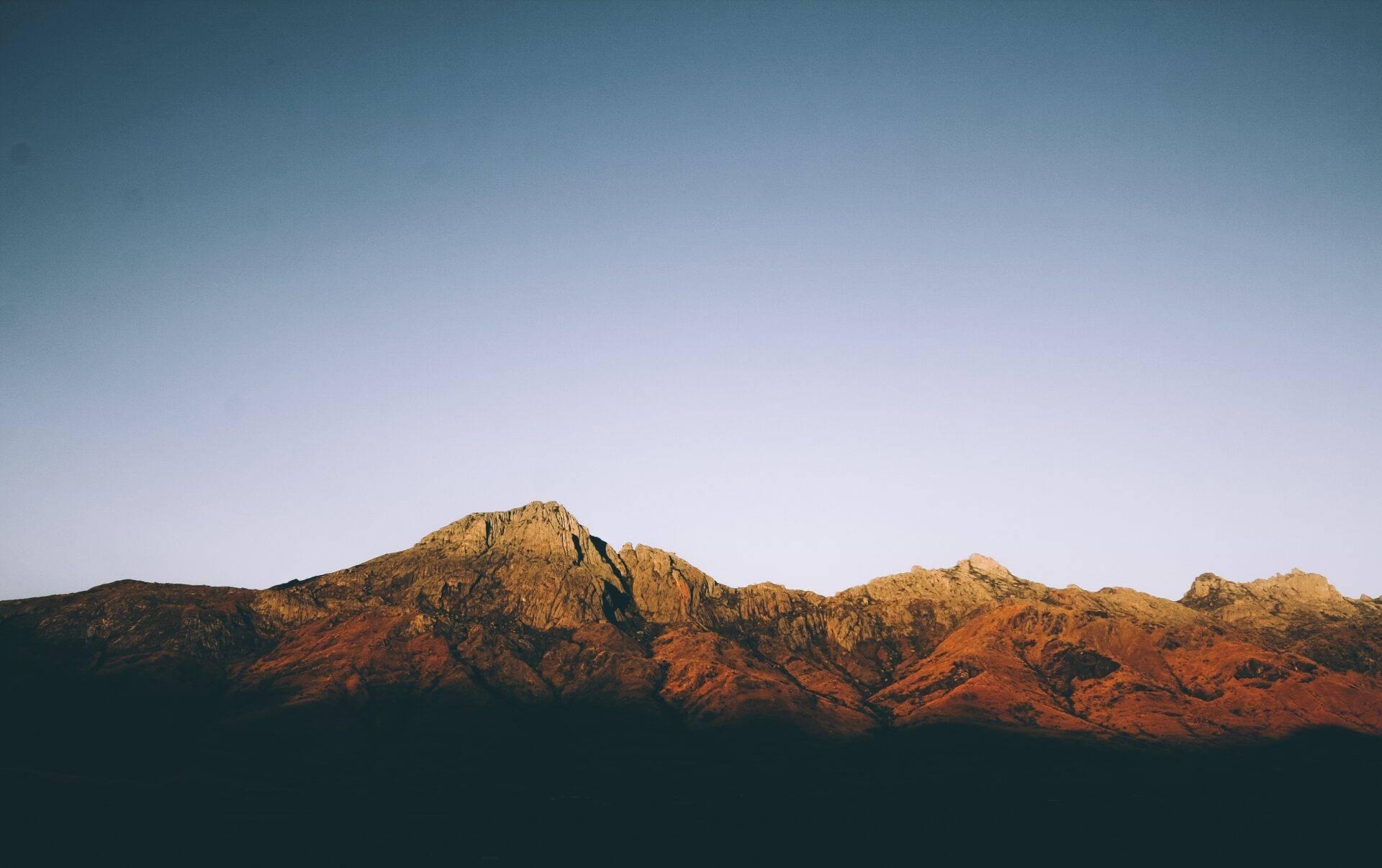 A rocky mountain ridge in warm ochre and reddish hues lit by the setting sun under a clear sky