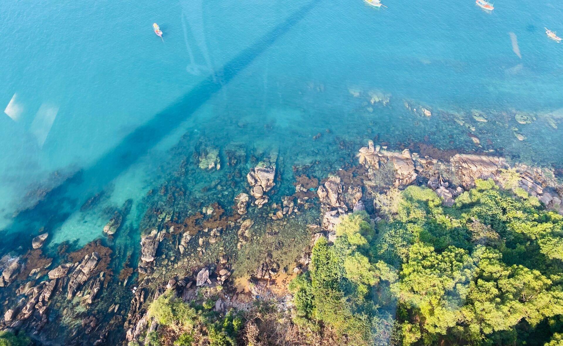 Vue aérienne d’une baie turquoise avec roches côtières, végétation verte et bateaux de pêche amarrés