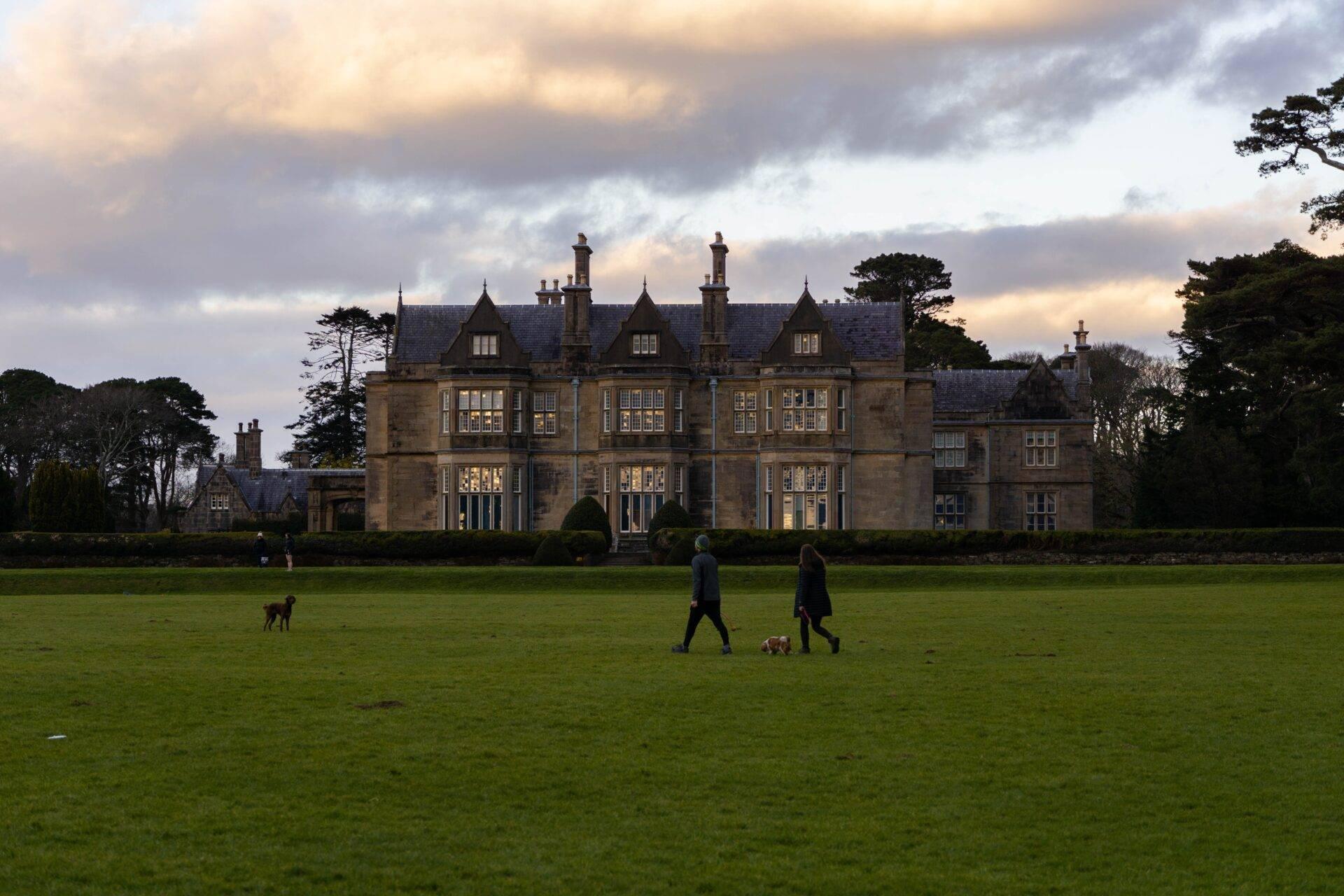 A large stone manor house with turrets, surrounded by a green park with people strolling