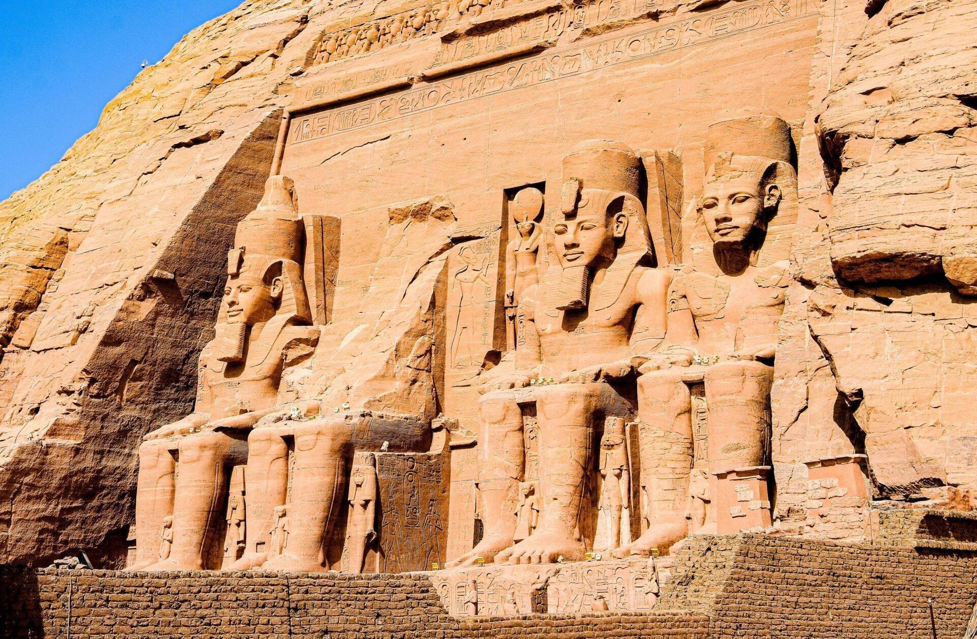 Rock-cut facade of an ancient Egyptian temple with four sculpted colossi and hieroglyphs beneath a blue sky