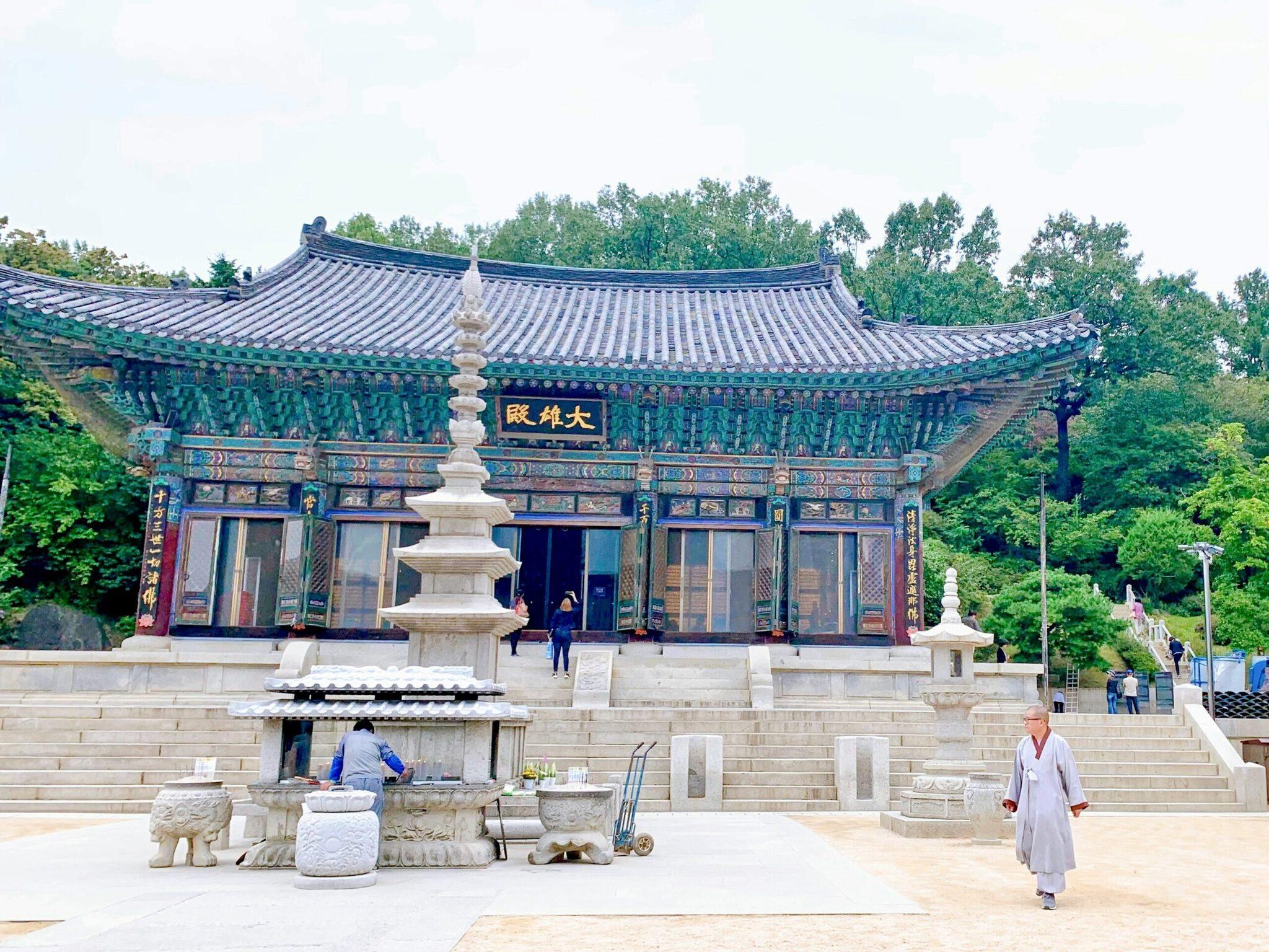 A Korean Buddhist temple with traditional blue and green tiled roof, a stone stupa, and visitors in a paved courtyard