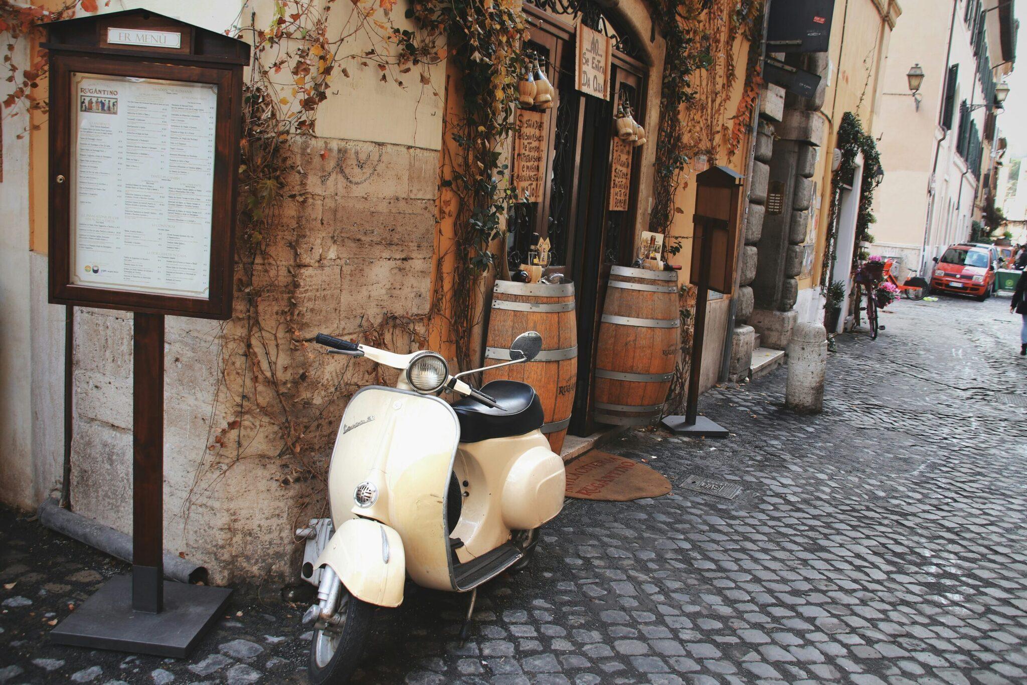 A white Vespa scooter parked in a cobbled alleyway of an old European city, with ochre facades and climbing ivy