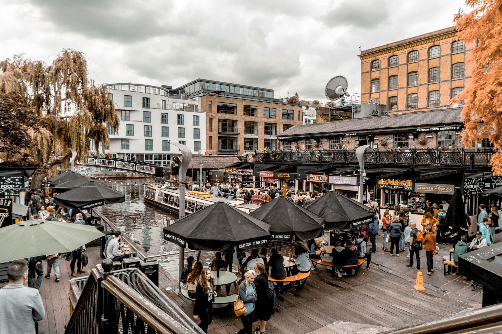 Un marché animé au bord d’un canal avec terrasses, boutiques et bâtiments modernes en arrière-plan par temps nuageux