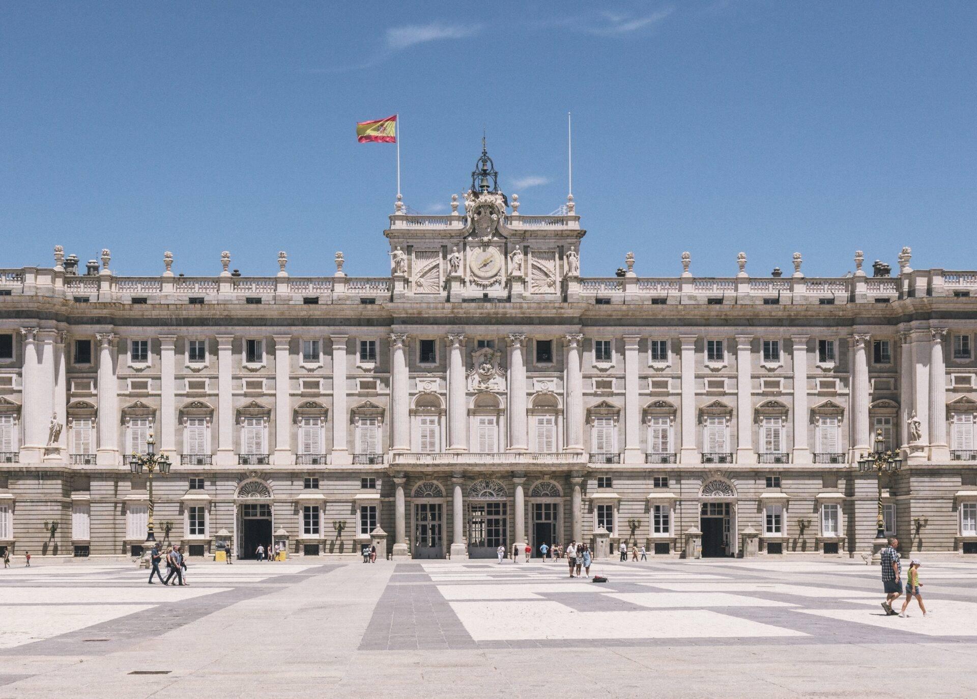 Façade majestueuse d’un palais blanc avec colonnes, horloge centrale et drapeau espagnol, cour pavée ensoleillée