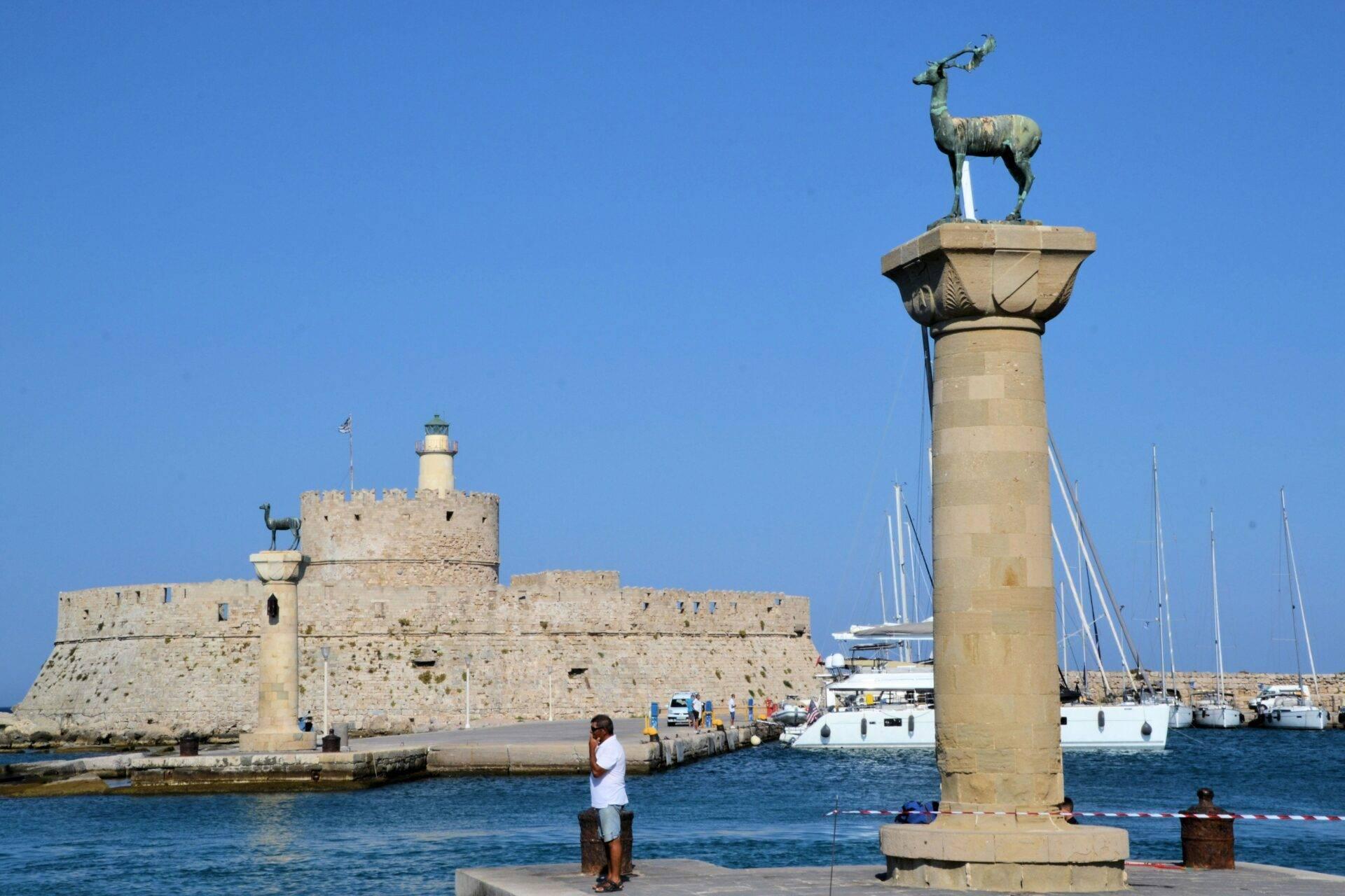 A Mediterranean harbour with a column topped by a stag statue, a stone fortress and sailing boats moored in the foreground