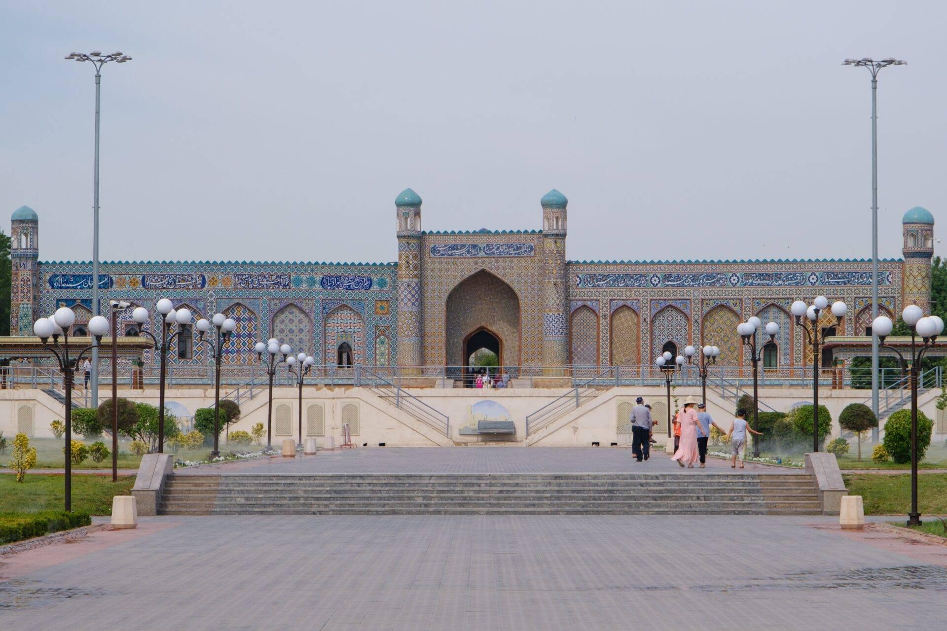 Large mosque with a blue and gold ornamental facade, central minaret, paved courtyard and visitors in light clothing