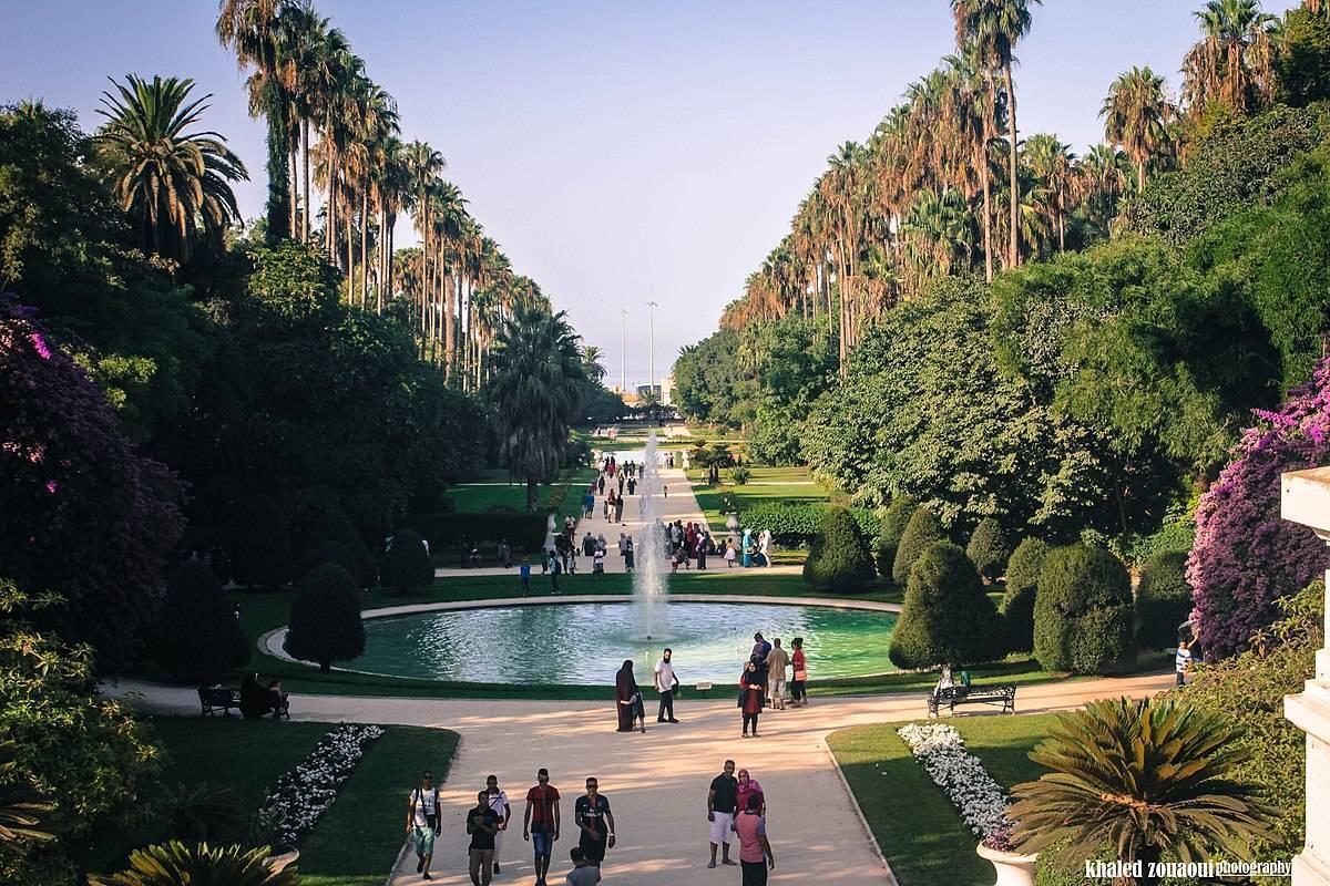 Un parc paysager avec fontaine centrale, allées bordées de palmiers et cyprès, verdure luxuriante et visiteurs se promenant