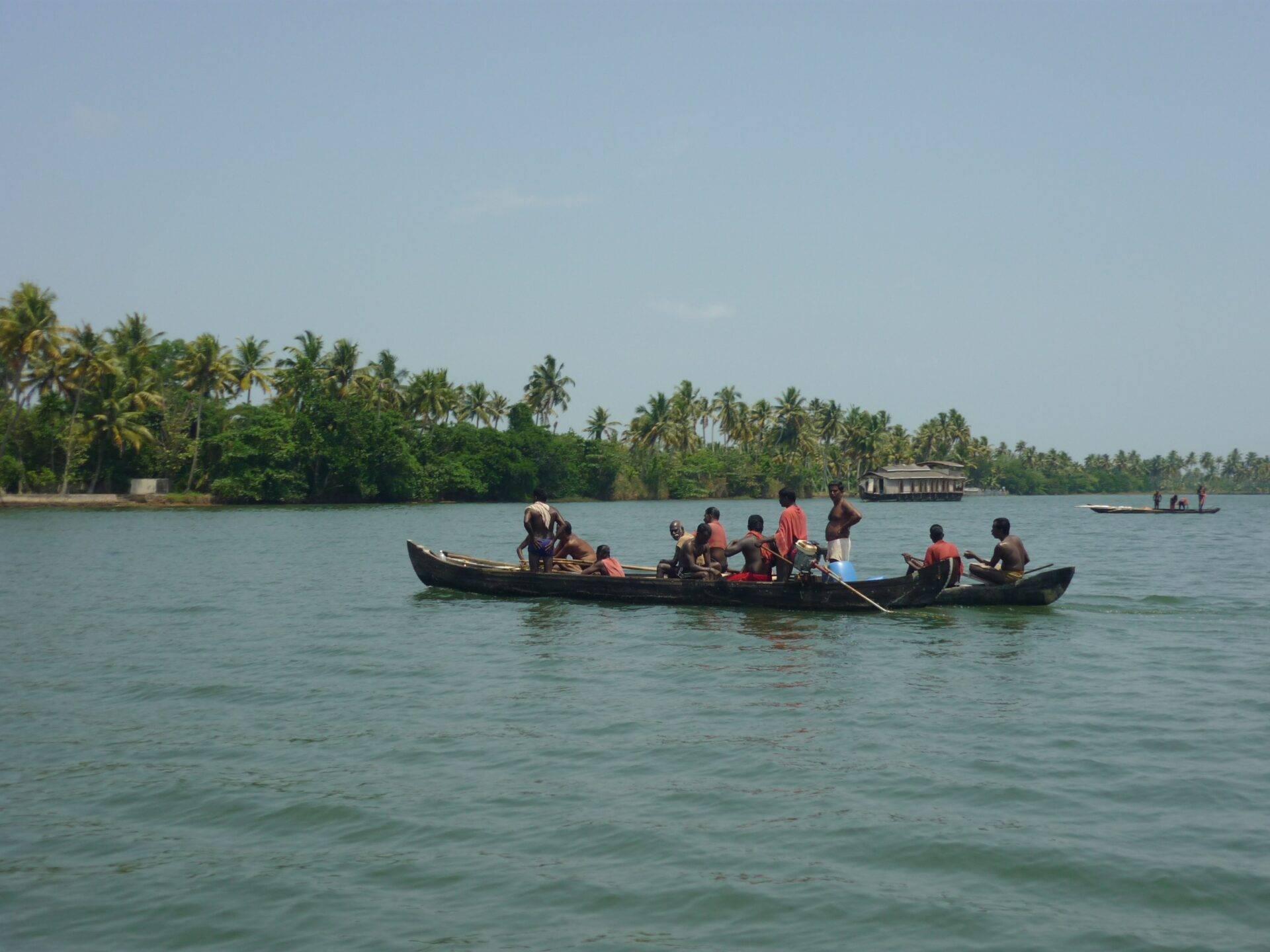 A traditional black boat glides across turquoise waters with palm trees and floating homes in the background
