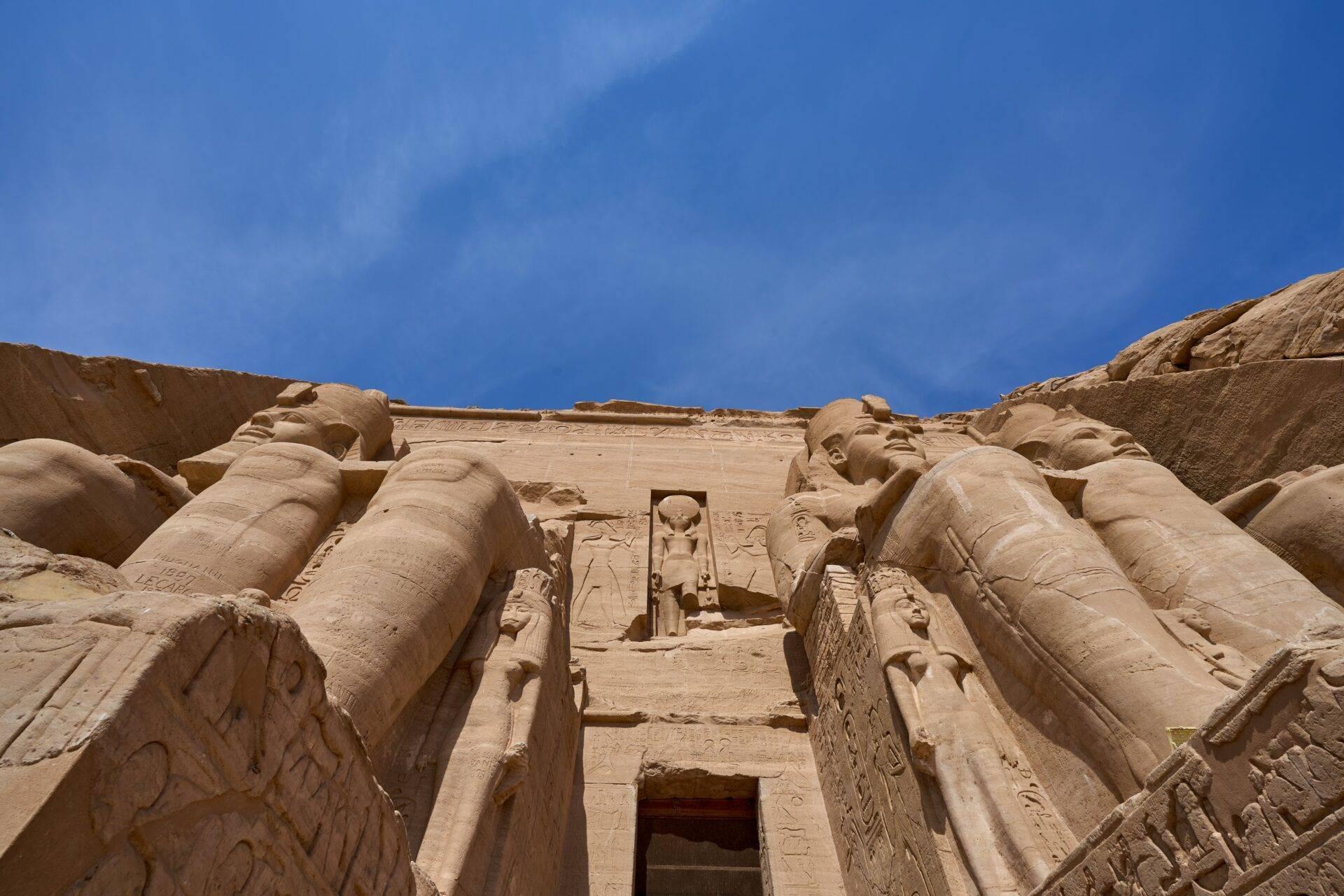 Monumental facade of an ancient Egyptian temple with sculpted colossi and hieroglyphs beneath a blue sky