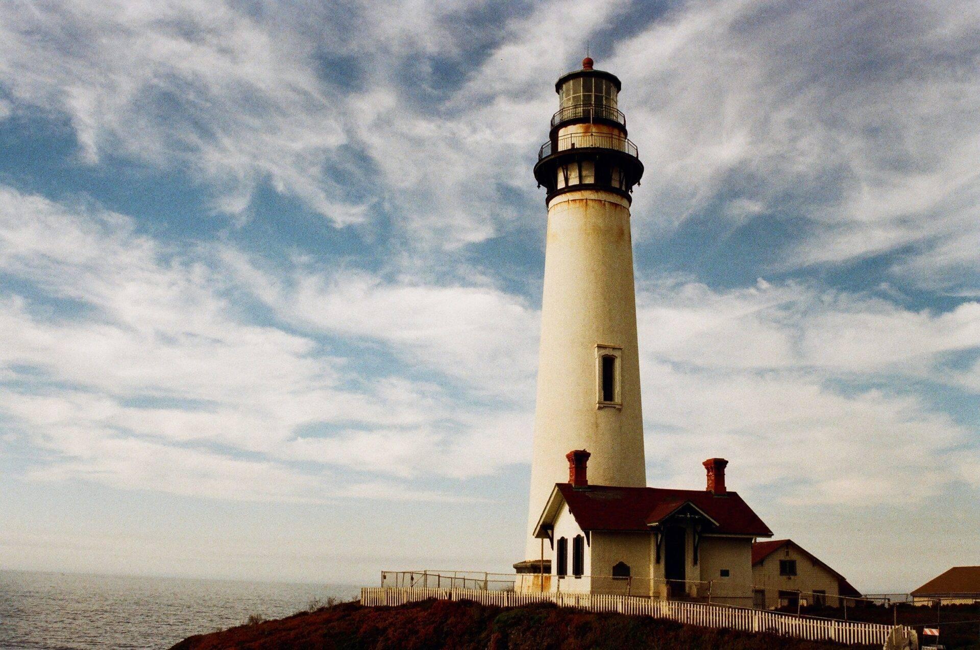 A white coastal lighthouse with its lantern room and red-roofed buildings under an overcast sky