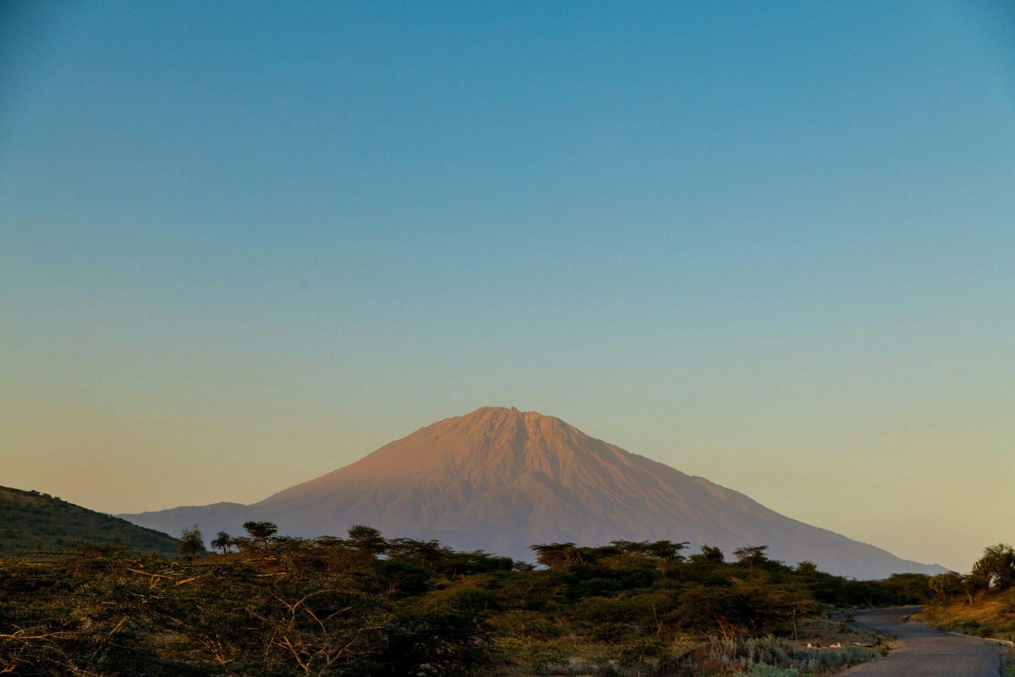 Un volcan majestueux aux pentes striées domine un paysage de savane aride avec acacias épars et une rivière en premier plan