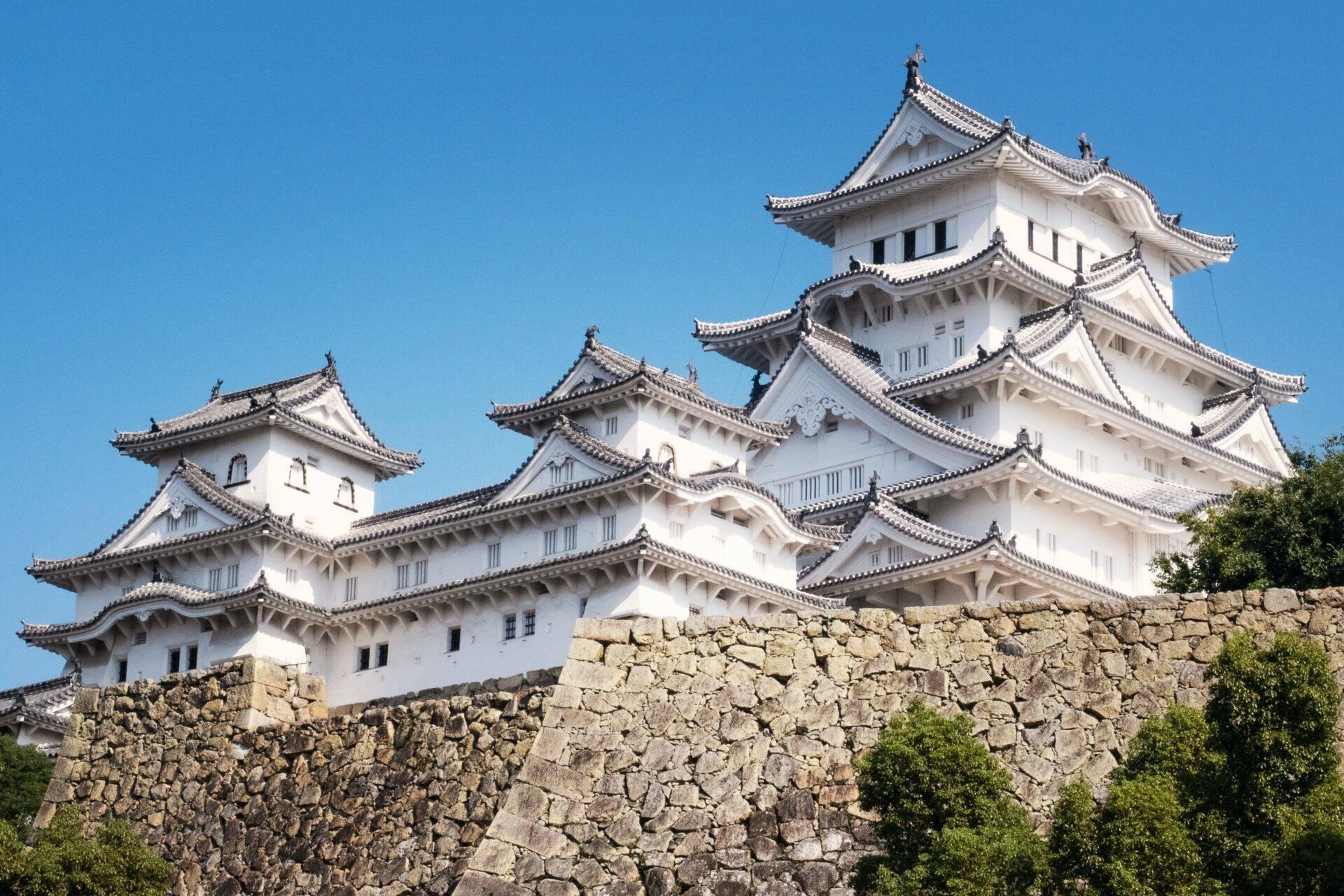 A white Japanese castle with curved rooflines and black roof tiles set on a stone wall beneath a blue sky