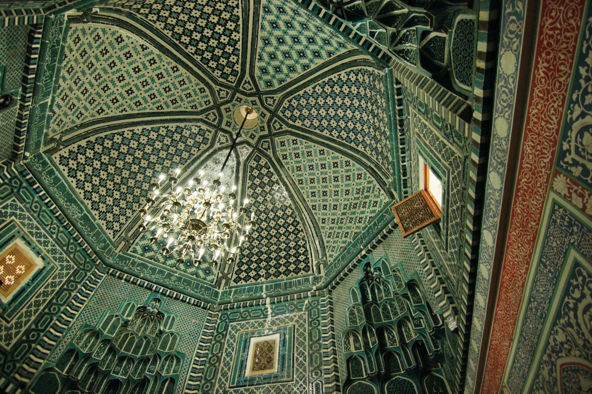 Aerial view of a mosque interior with an ornate octagonal ceiling, turquoise tilework and a central chandelier