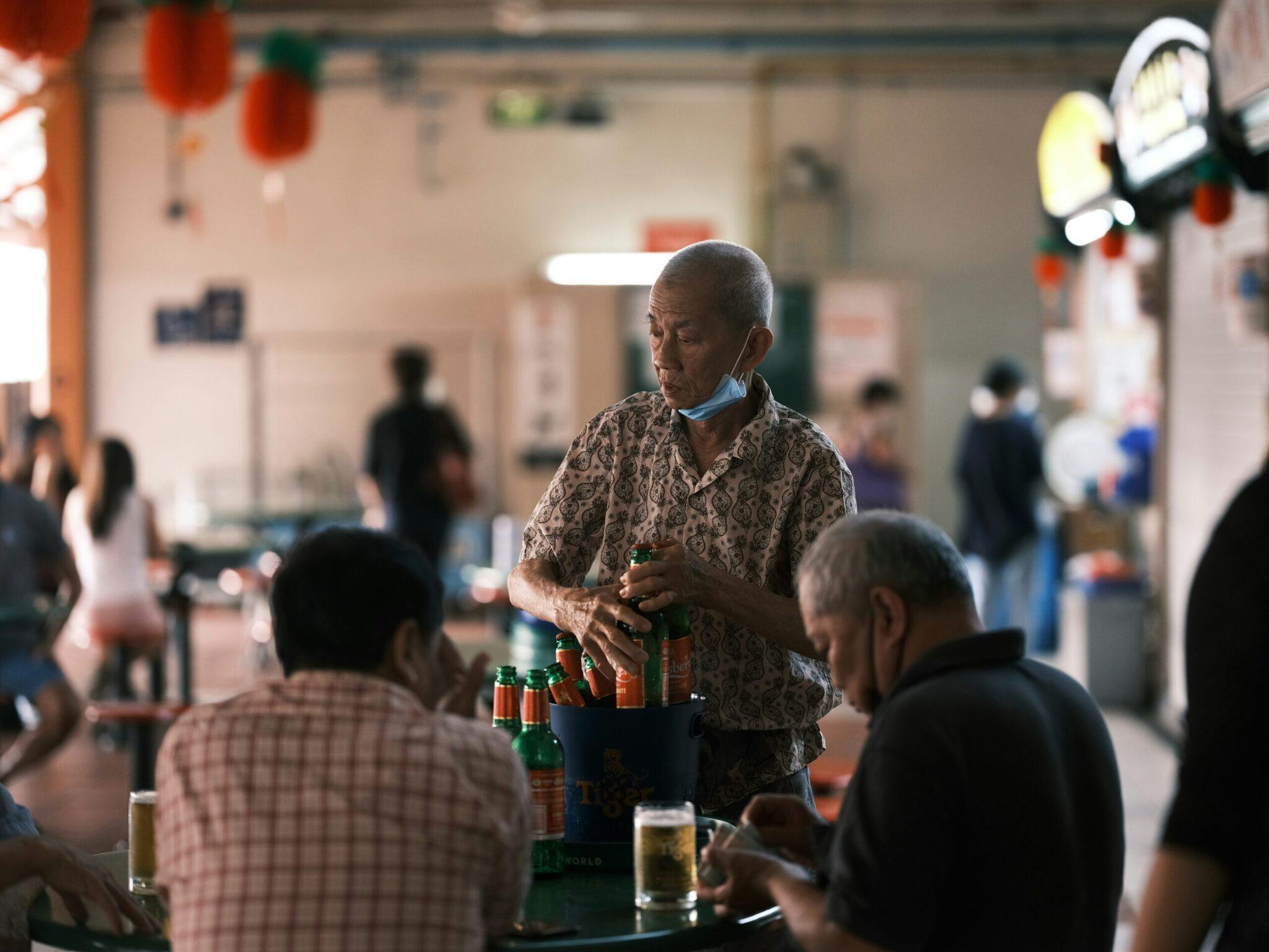 Un homme âgé servant des bières lors d’une célébration communautaire dans un hall décoré de lanternes rouges